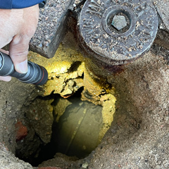 A worker uses a flashlight to examine a water service line visible within a pothole.