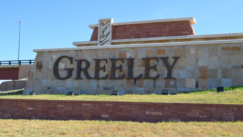 A prominent stone and brick welcome sign displaying 'GREELEY' with metal lettering, situated near a highway overpass under a clear blue sky.