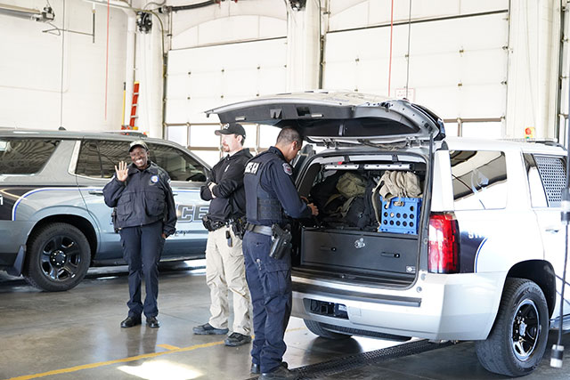Two policemen and a policewoman waving and inspecting equipment in police vehicles in a department garage