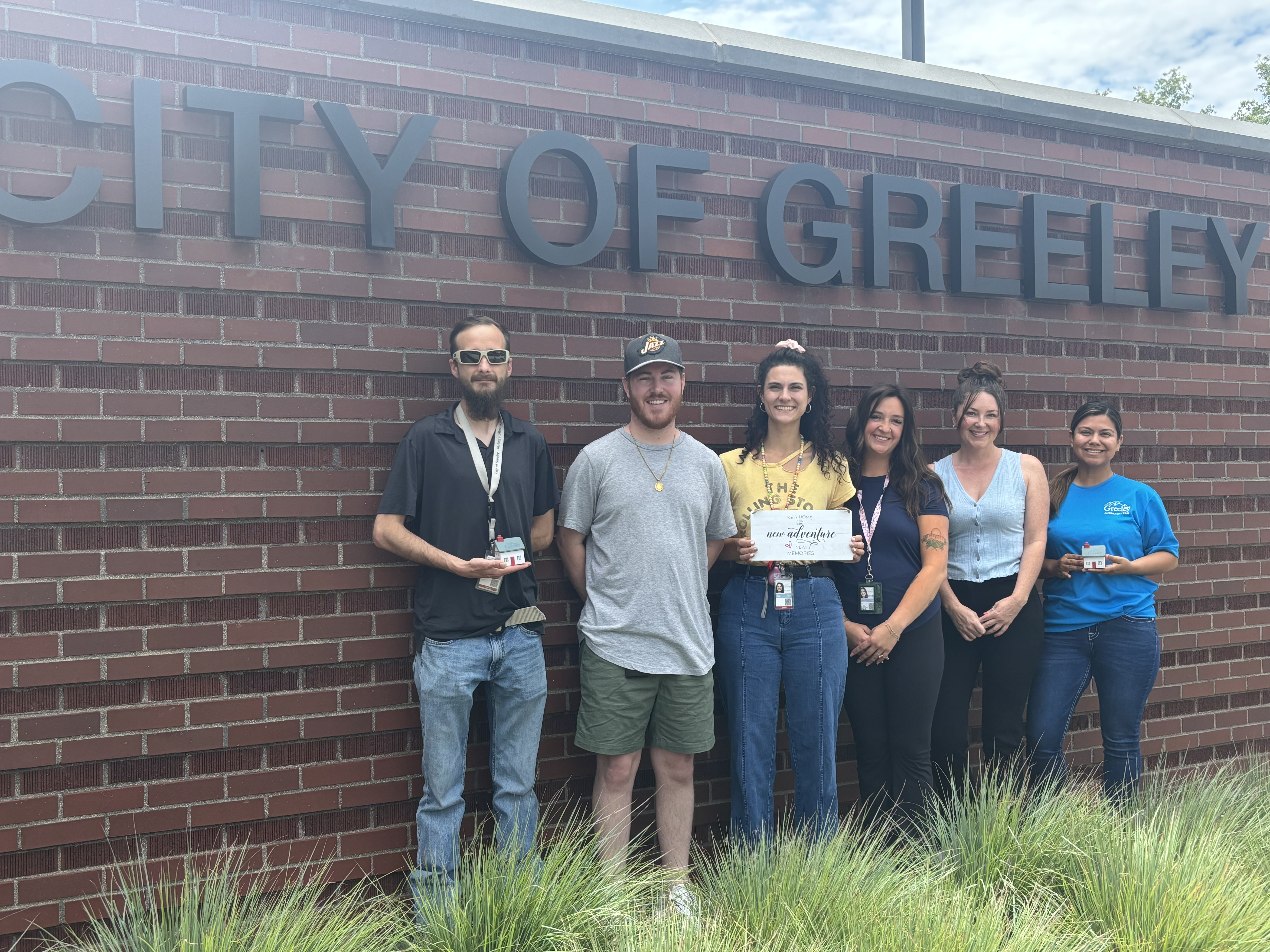 Six City of Greeley Outreach employees stand in front of a brick wall with the words "City of Greeley." Three individuals hold recognition items, including a sign labeled "new home, new adventure" and two small model houses. Tall grasses are in the foreground.