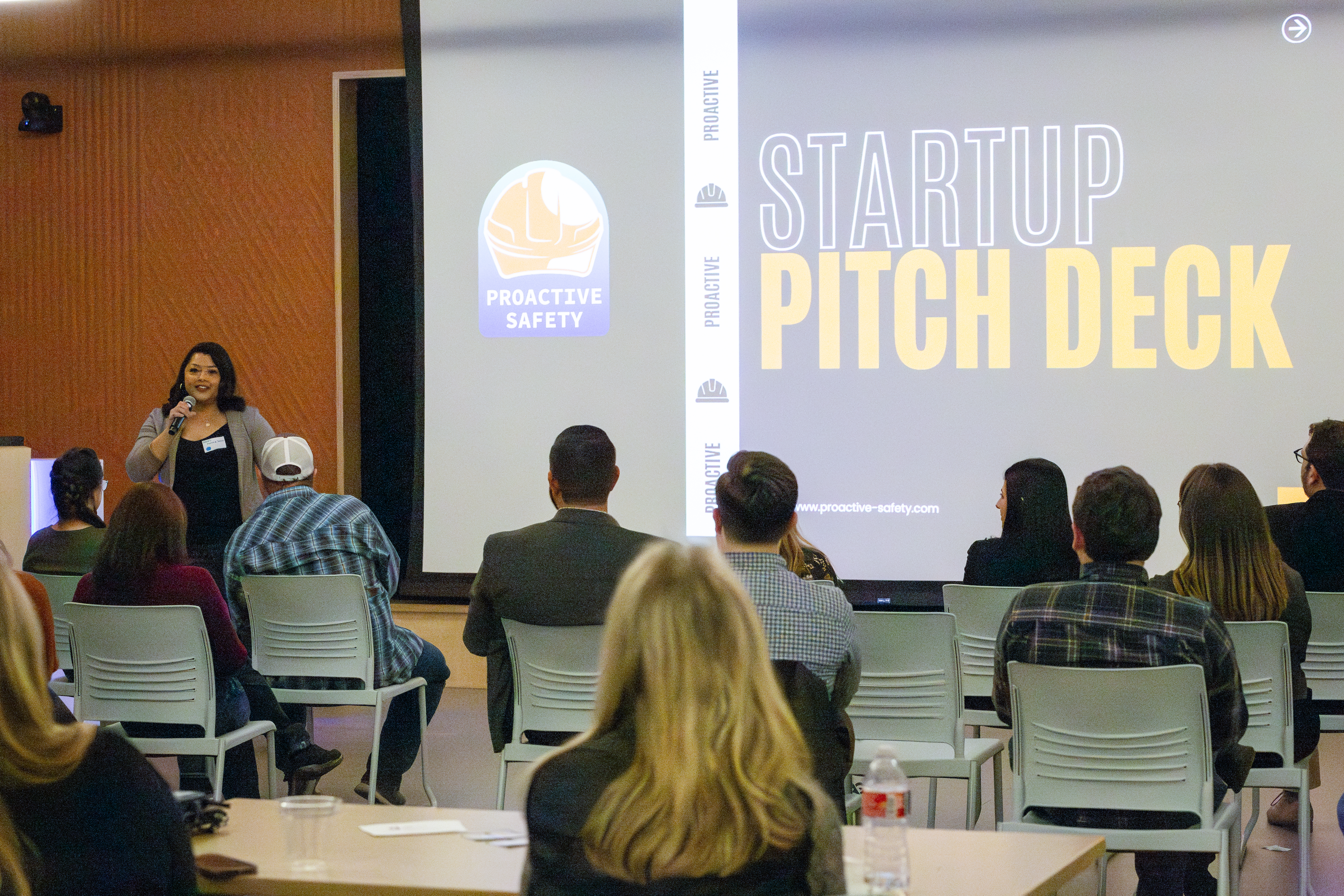This image depicts a gBETA meeting where Greeley entrepreneurs pitch their business ideas to investors and other business resources.  A woman is standing at the front of the room holding a microphone and behind her, a presentation slide displays the words "STARTUP PITCH DECK".  Audience members watch from rows of chairs in front of the speaker.