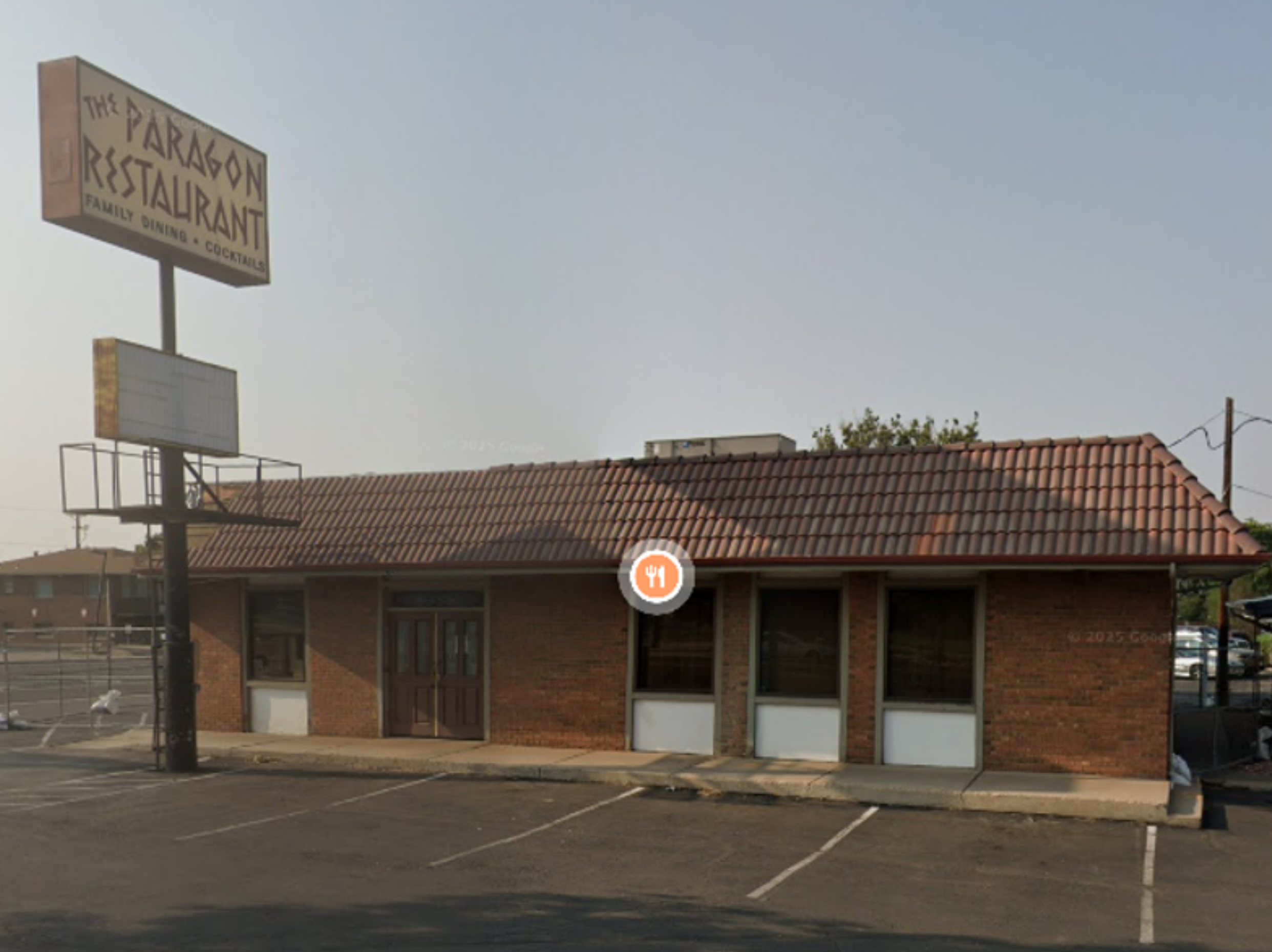 This image depicts the Paragon Family Restaurant in Greeley, Colorado, before a facade improvement project. A dated brown brick building with a red clay tile roof sits behind a pole sign displaying the name of the restaurant in Greek style lettering.  