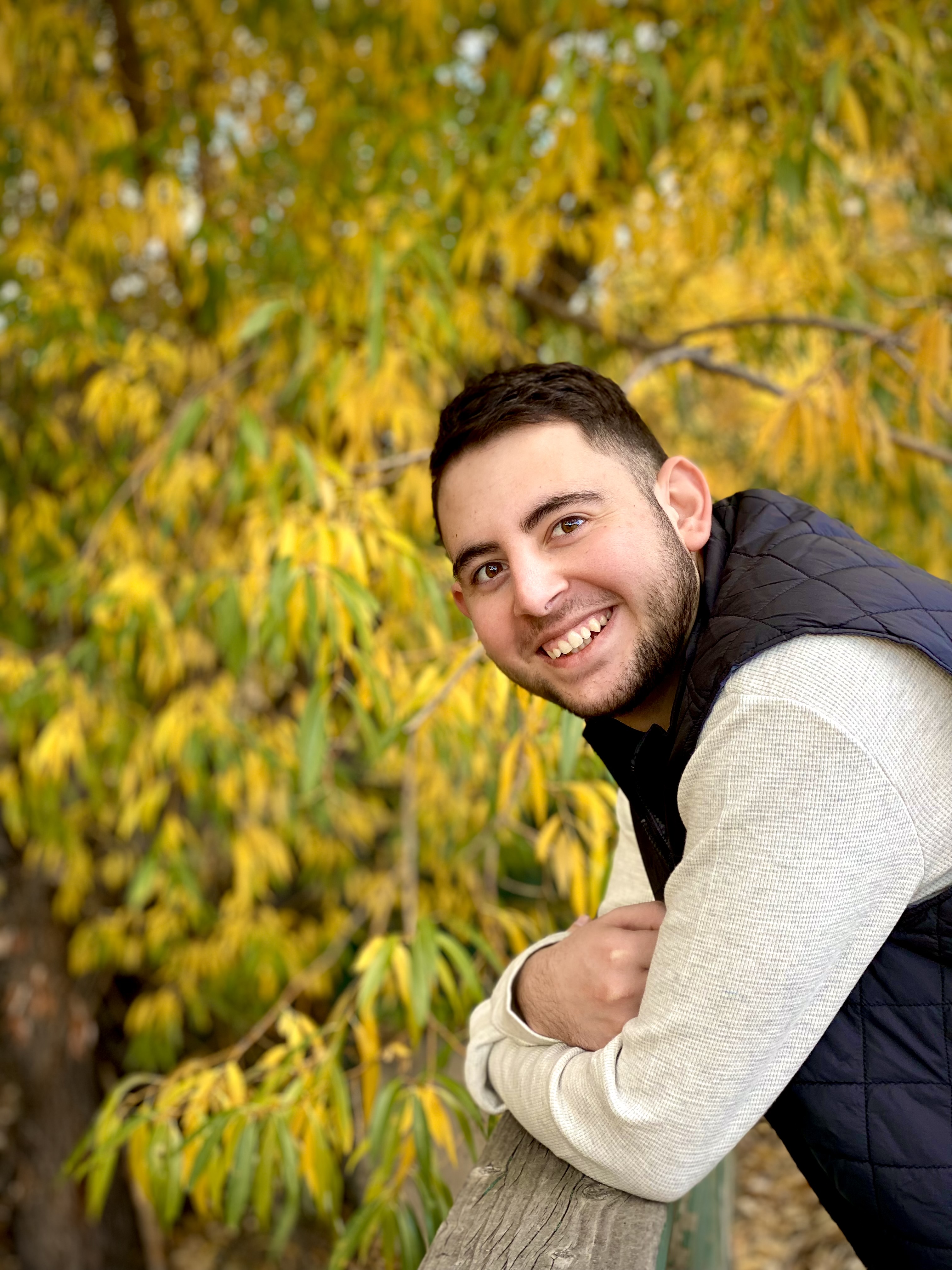 Photo of GPD Co-Responder Basal Hamdan. He is wearing a beige long-sleeved shirt with a black vest. He is leaning on a wood railing with yellow and green leaves out of focus in the background.