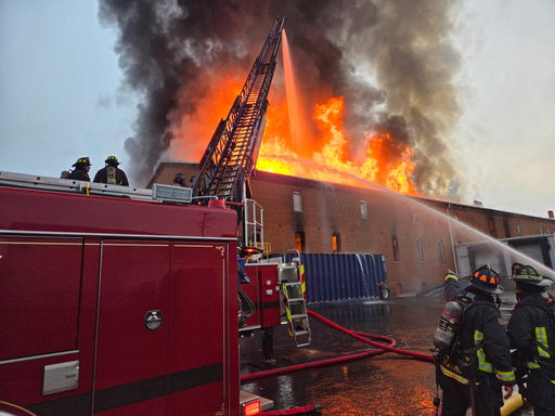 5 Greeley firefighters are pictured on the scene of a large building fire. A ladder truck is spraying water onto the fire and a hose stream can also been seen spraying the fire. 