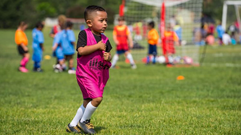 Young boy walks wearing a sports penny, while other children are in the background playing soccer, on the park field during a youth sports soccer camp