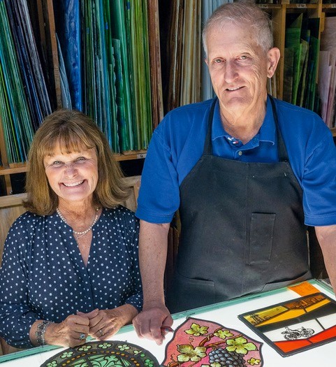 Watkins couple standing in their stained glass workshop smiling at the camera