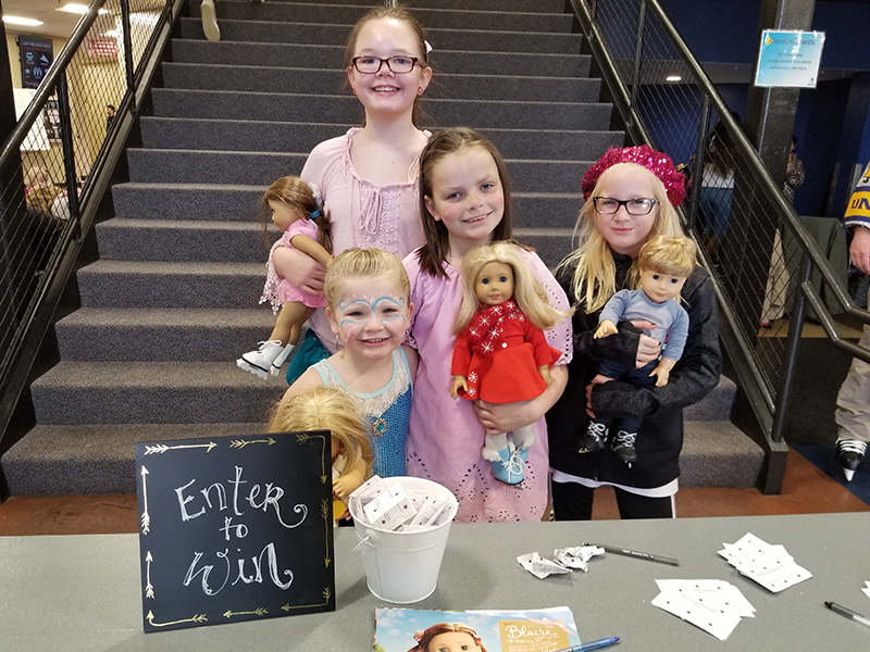Four girls smile holding their American Girl Dolls by a raffle cup that reads "enter to Win" at the Greeley Ice Haus
