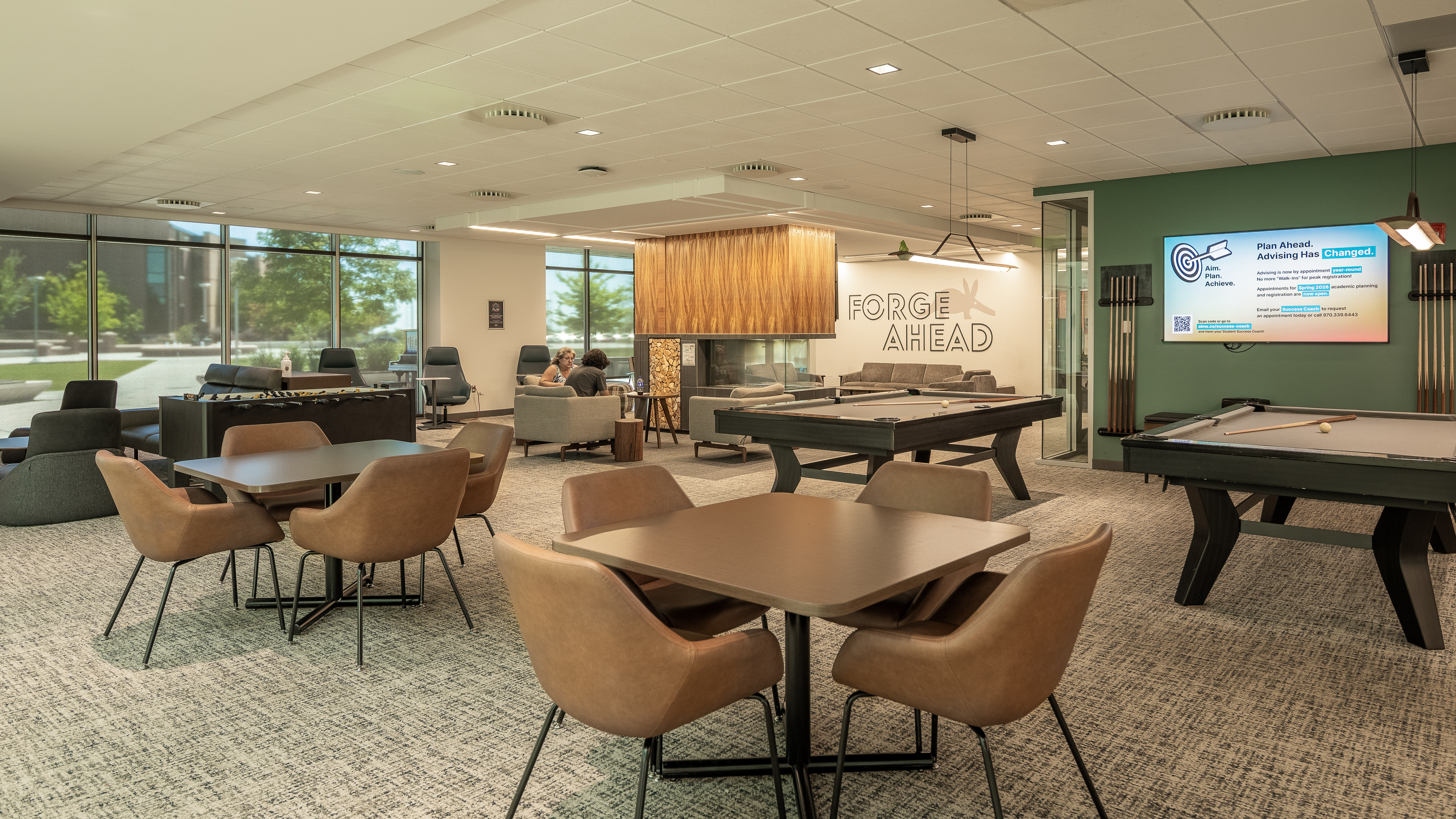 Interior student lounge at Aims Community College with seating areas, game tables, large windows, and a “Forge Ahead” wall graphic.