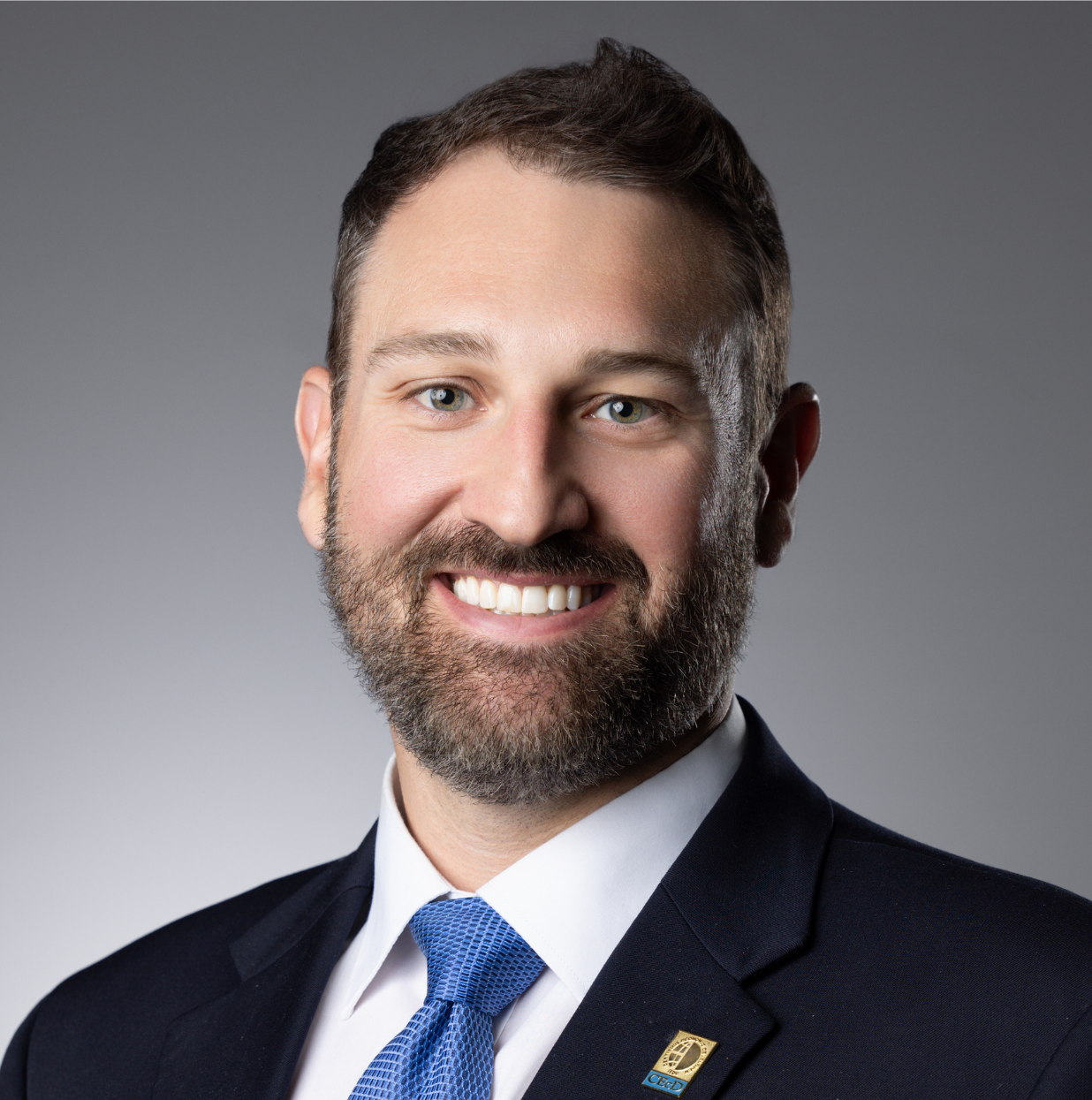 This image depicts a professional headshot of Economic Development Manager Derrek Jerred.  Derrek is shown in a dark suit jacket, white dress shirt, and blue tie.  