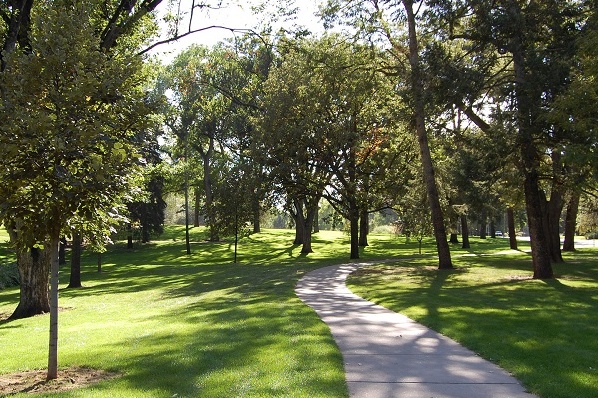 A serene park scene with a paved walkway winding through lush greenery and mature trees.