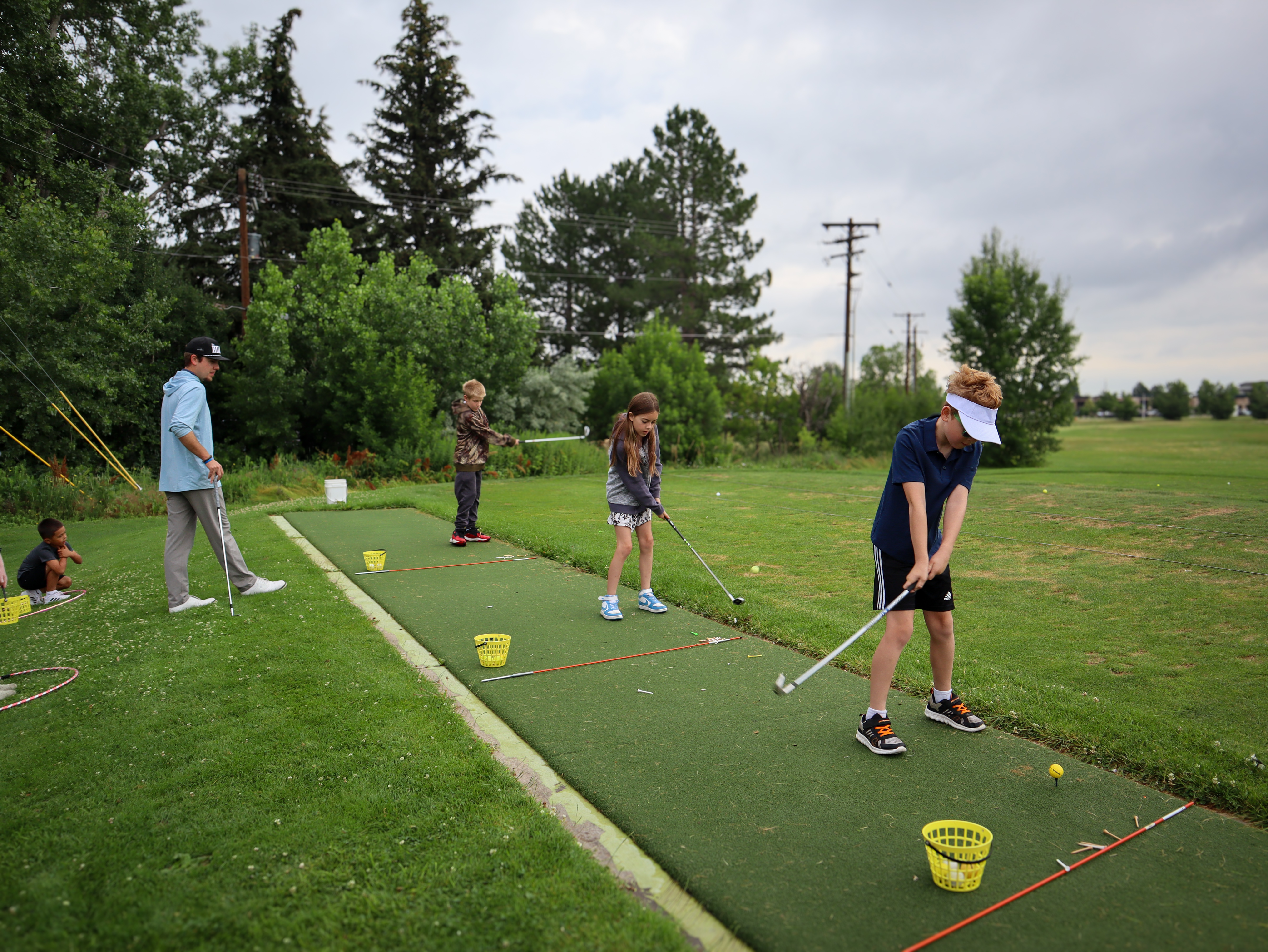 Children practicing golf swings on green mats at an outdoor driving range with an instructor nearby observing.