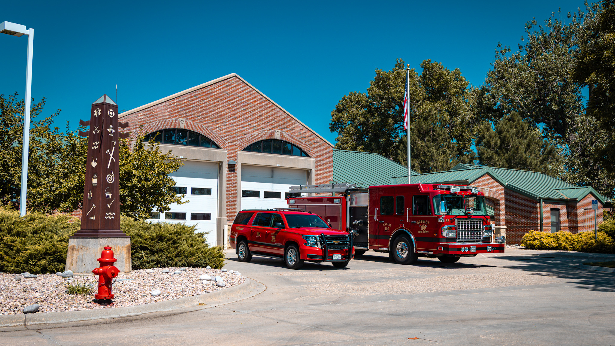 A picture showing a fire station with two fire trucks staged out in front of it. The fire station can be seen behind the trucks with a flag flying in the background. 