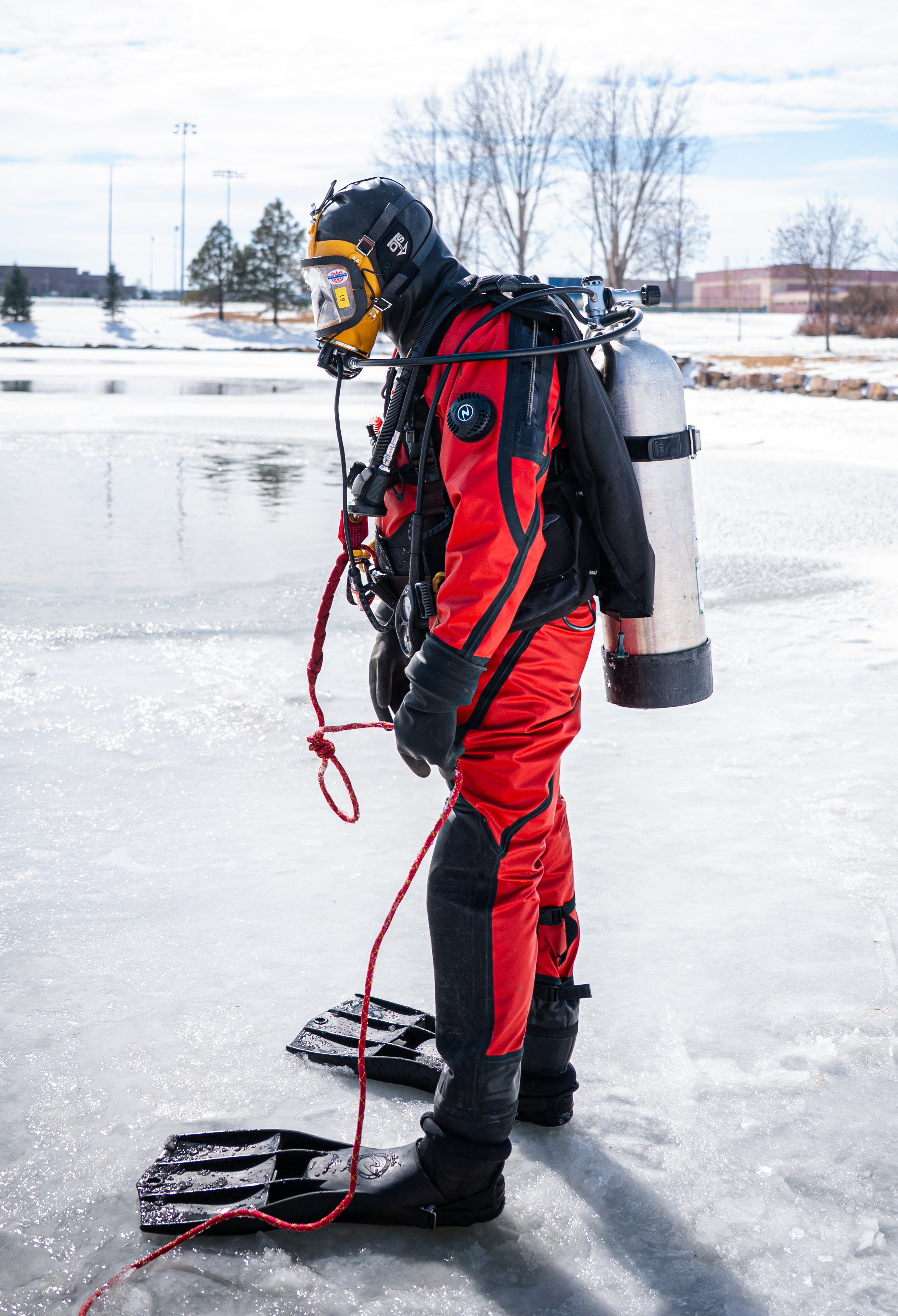 A Greeley firefighter member of the Dive Rescue Team in scuba gear and an orange wetsuit stands in front of a frozen pond with a residential neighborhood behind him daytime.