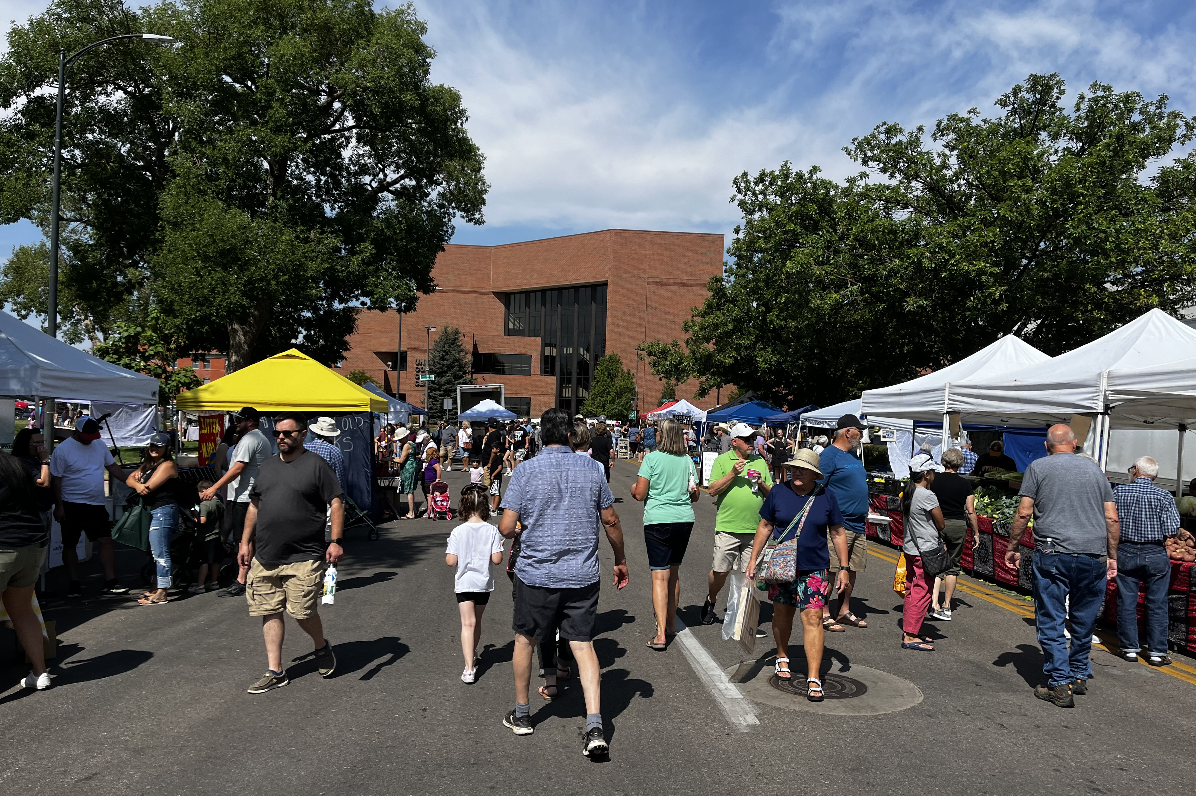 A bright morning at the Greeley Farmers' Market, boasts a crowd of many people along a tent lined street of vendors with the UCCC in the background. 