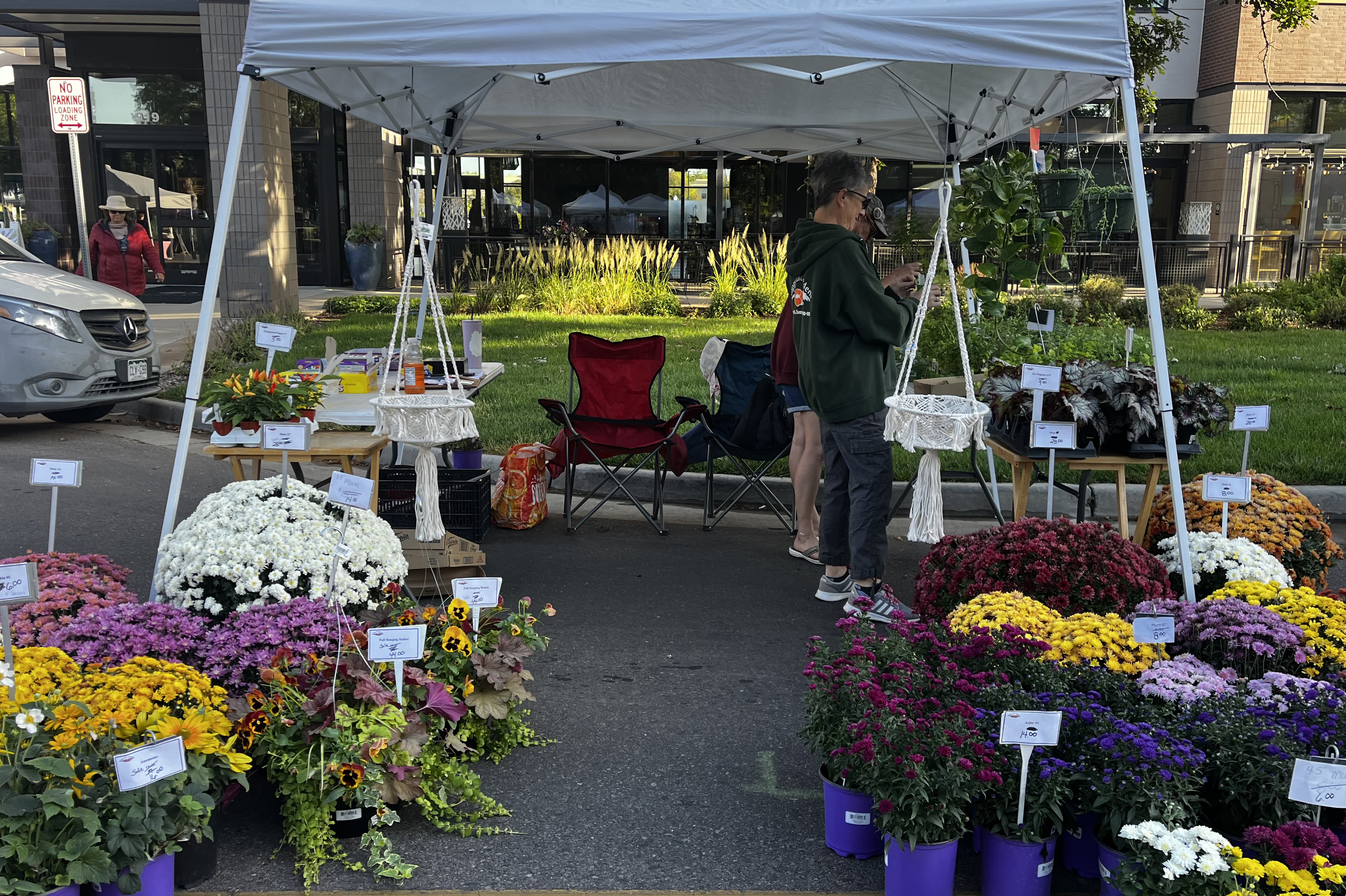 A booth full of flowers in bloom at the Greeley Farmers Market. Two people are under the booth tent, and the flowers, mostly bright Chrysanthemums in white, yellow, pink and red, extend out of the tent on either side. 