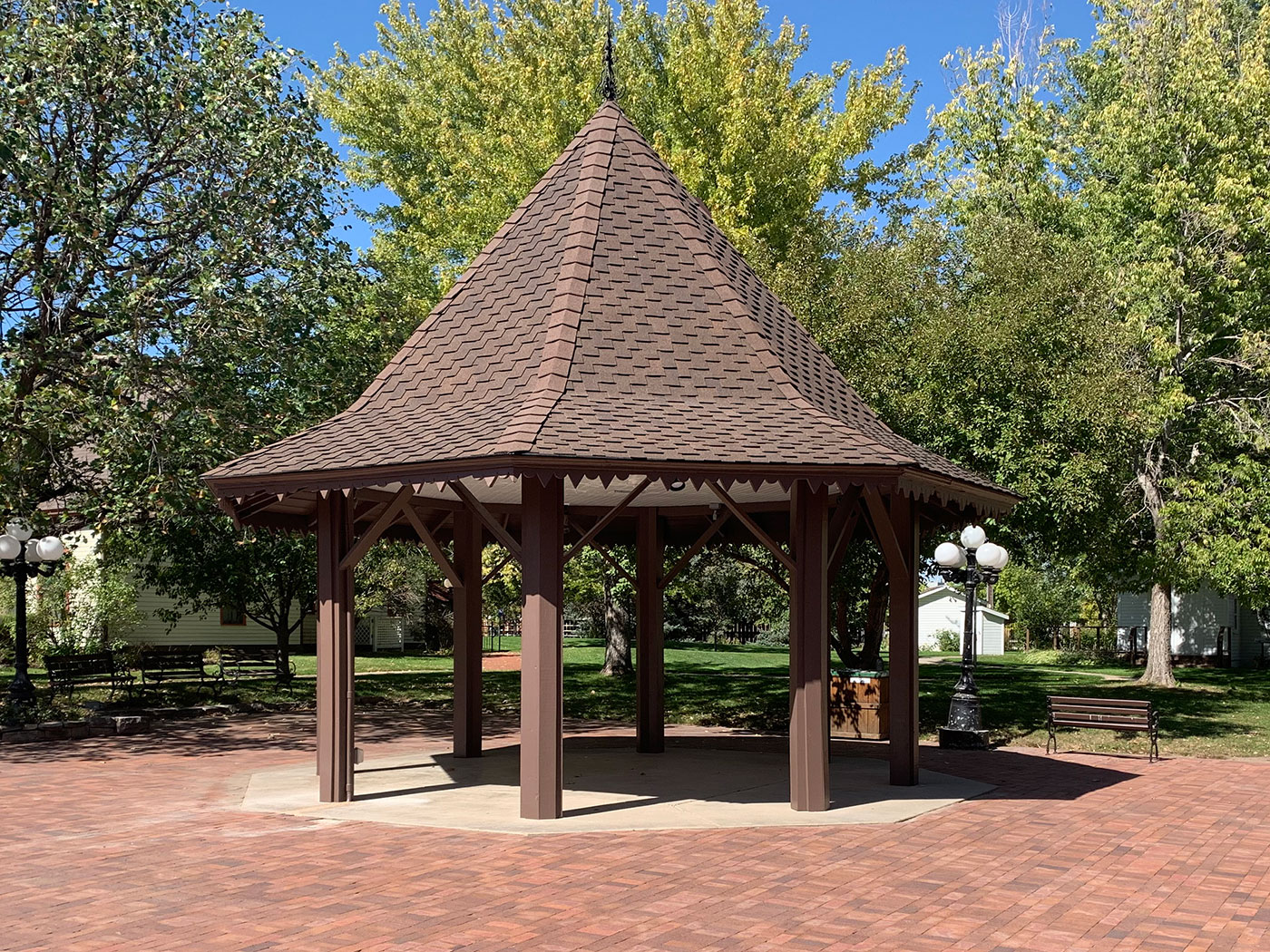 Polygonal gazebo with a steeply pitched roof and a brick patio in front of trees on a sunny summer day