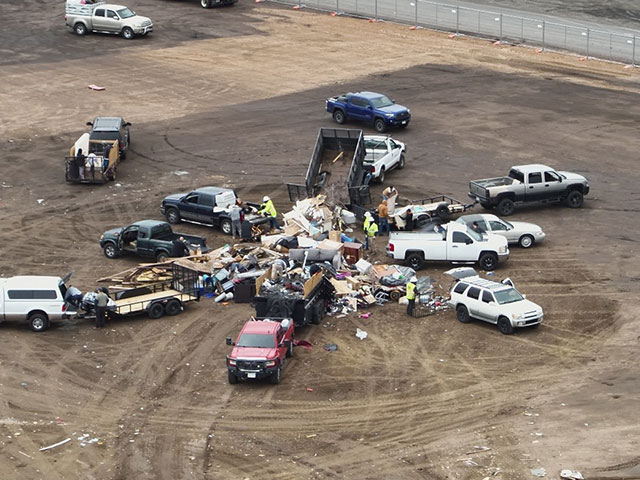 Cars and pickup trucks parked in reverse on a dirt lot around a sizeable garbage heap. Workers in safety vests and helmets help drivers safely unload their vehicles.