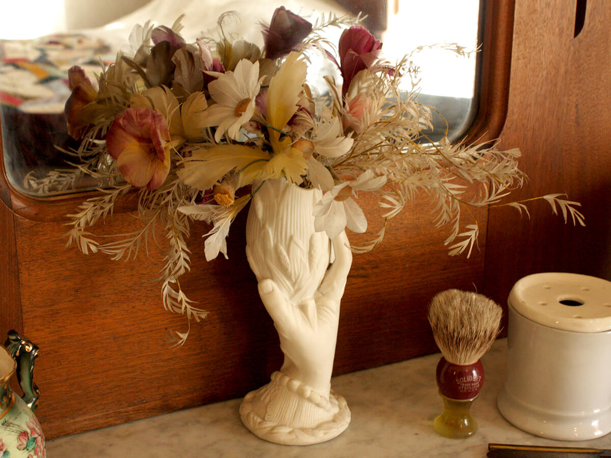 A sculptural vase resembling a hand holding a cup with stylized corn carvings contains dried flowers and sits on a countertop in front of a mirror