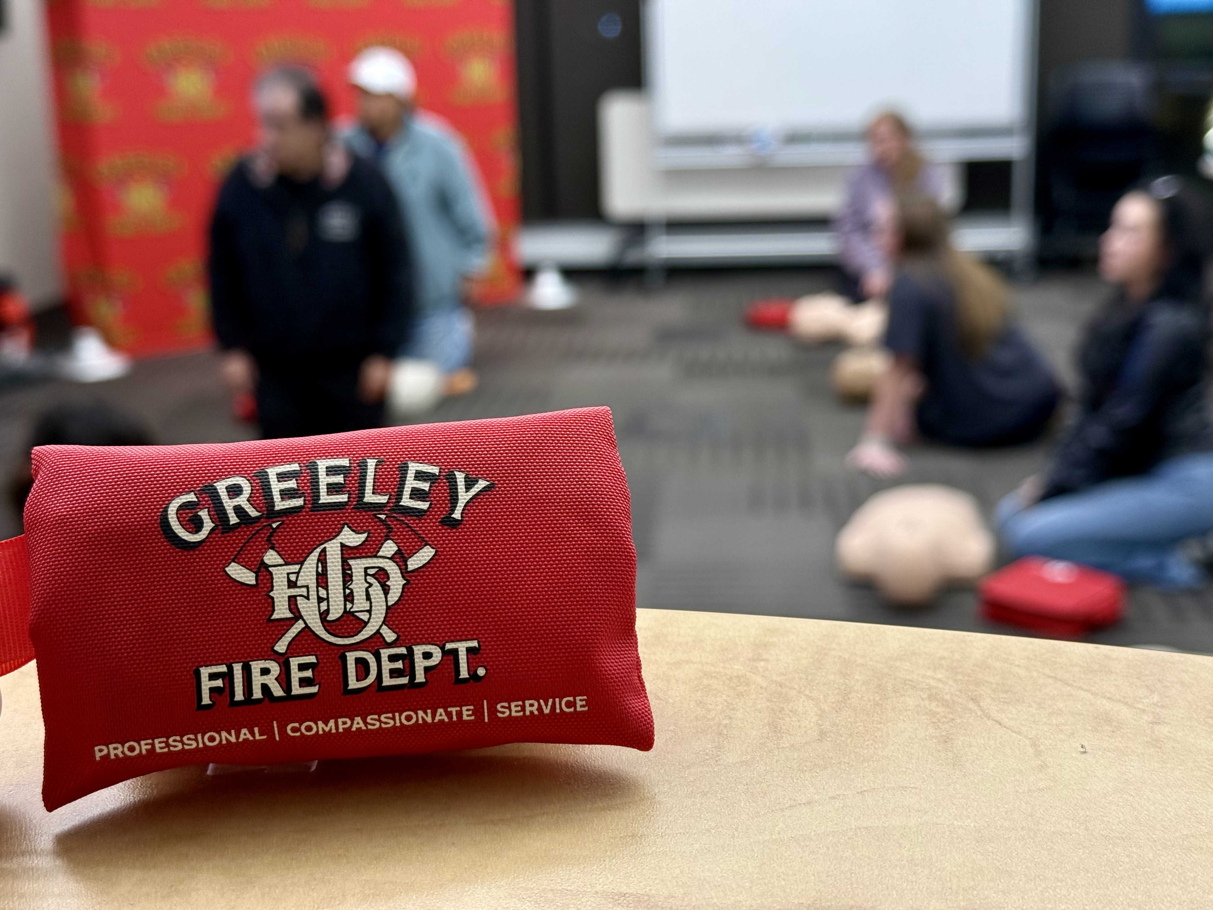 A keychain pack of CPR supplies with the words "Greeley Fire Department; Professional; Compassionate; Service" sits on a table in front of a blurred shot of members of the public attending a CPR class.