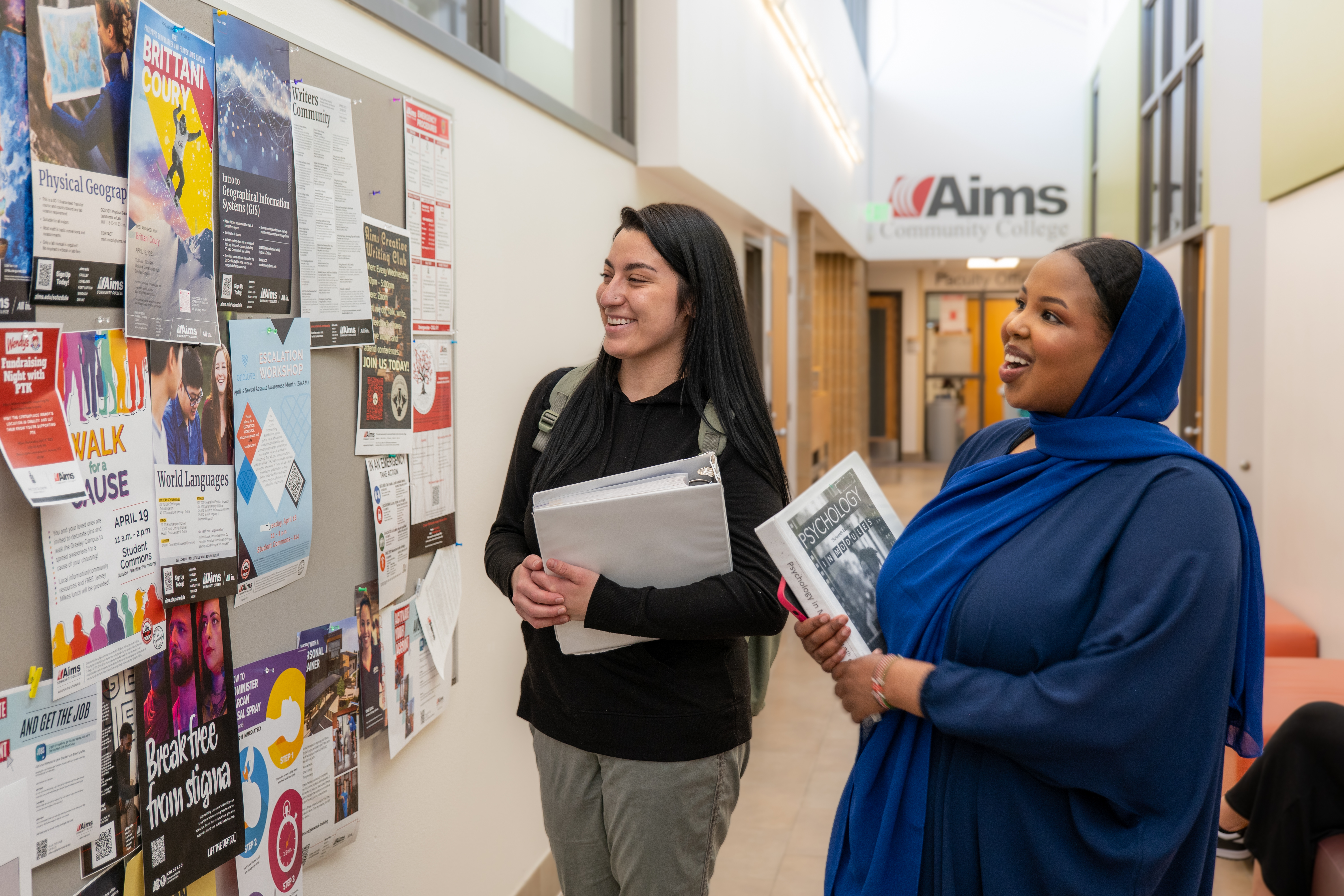 Two women are smiling and standing in a hallway reading bulletins pinned to a cork board.  The woman on the left has long, dark hair and is wearing a black sweatshirt and gray pants.  The woman on the right is wearing a navy blue blouse and bright blue head scarf.  Both women hold white binders and the cover of one reads "Psychology".  Behind the women is another hallway with Aims Community College logo painted above the doorway.  