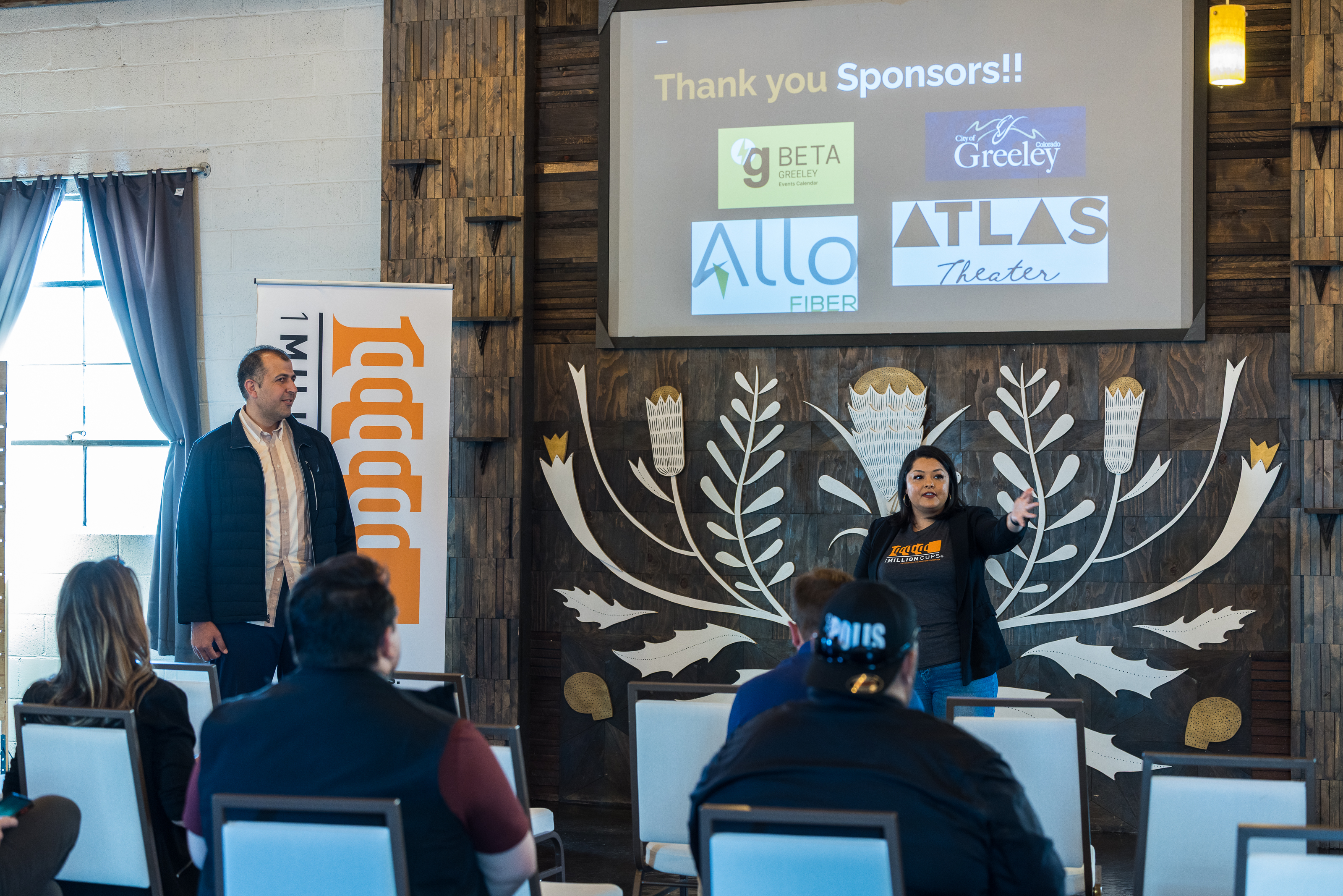 This image depicts a 1 Million Cups meeting where Greeley entrepreneurs network and obtain business training.  A man and woman are standing at the front of the room.  The woman's hand it outstretched, addressing an audience member's question.  Audience members watch from rows of chairs in front of the speakers.  A presentation screen behind the speakers displays business logos for the City of Greeley, gBETA, Allo Fiber, and Atlas Theater.