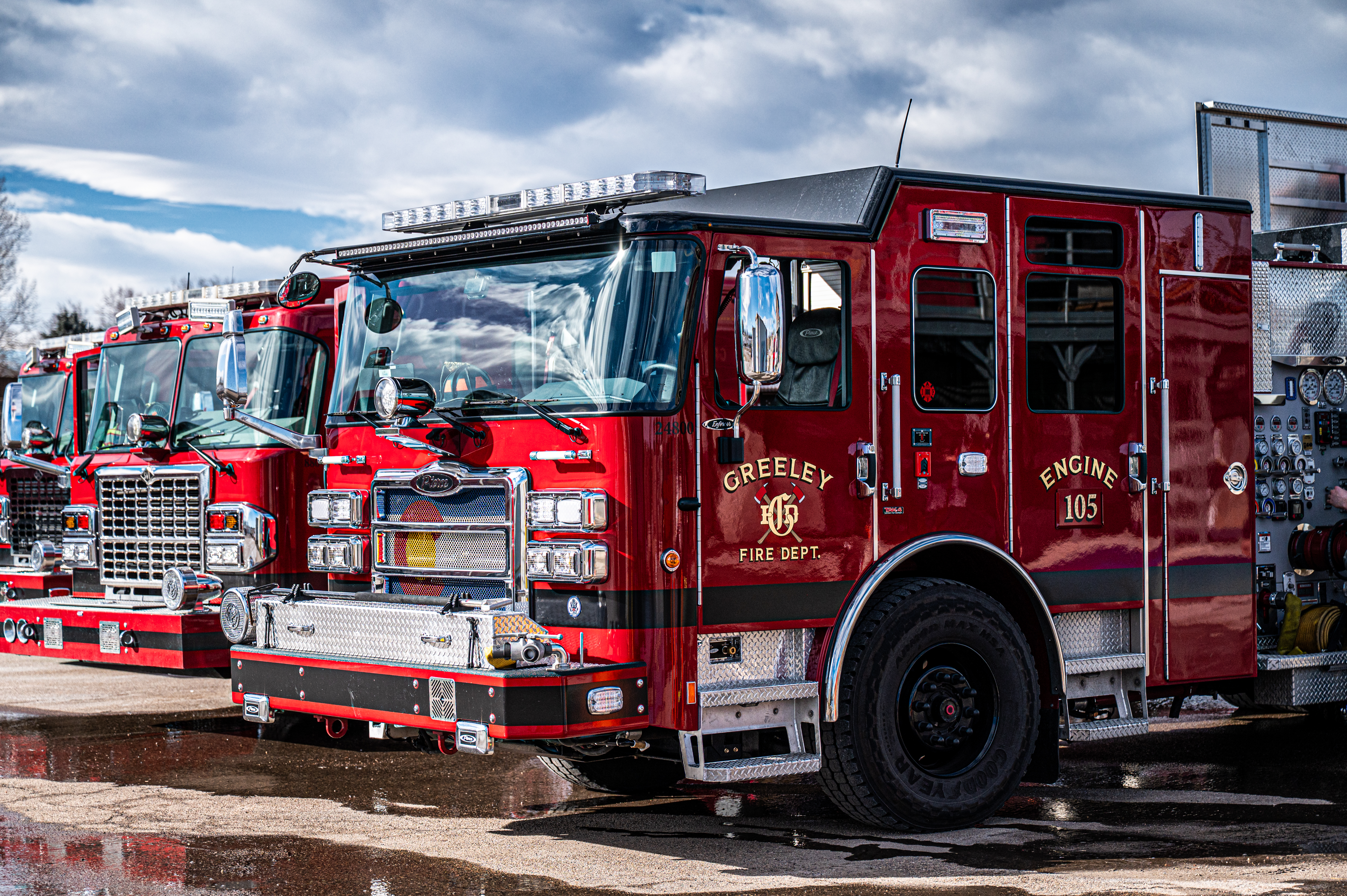Three front ends of red Greeley Fire Trucks parked side by side outdoors with water on the ground and a cloudy blue sky above