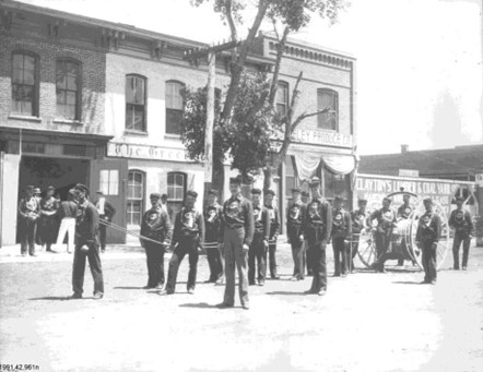 Historic image showcasing members of the Greeley Fire Department in uniform with equipment, standing in front of buildings from the 1880s.