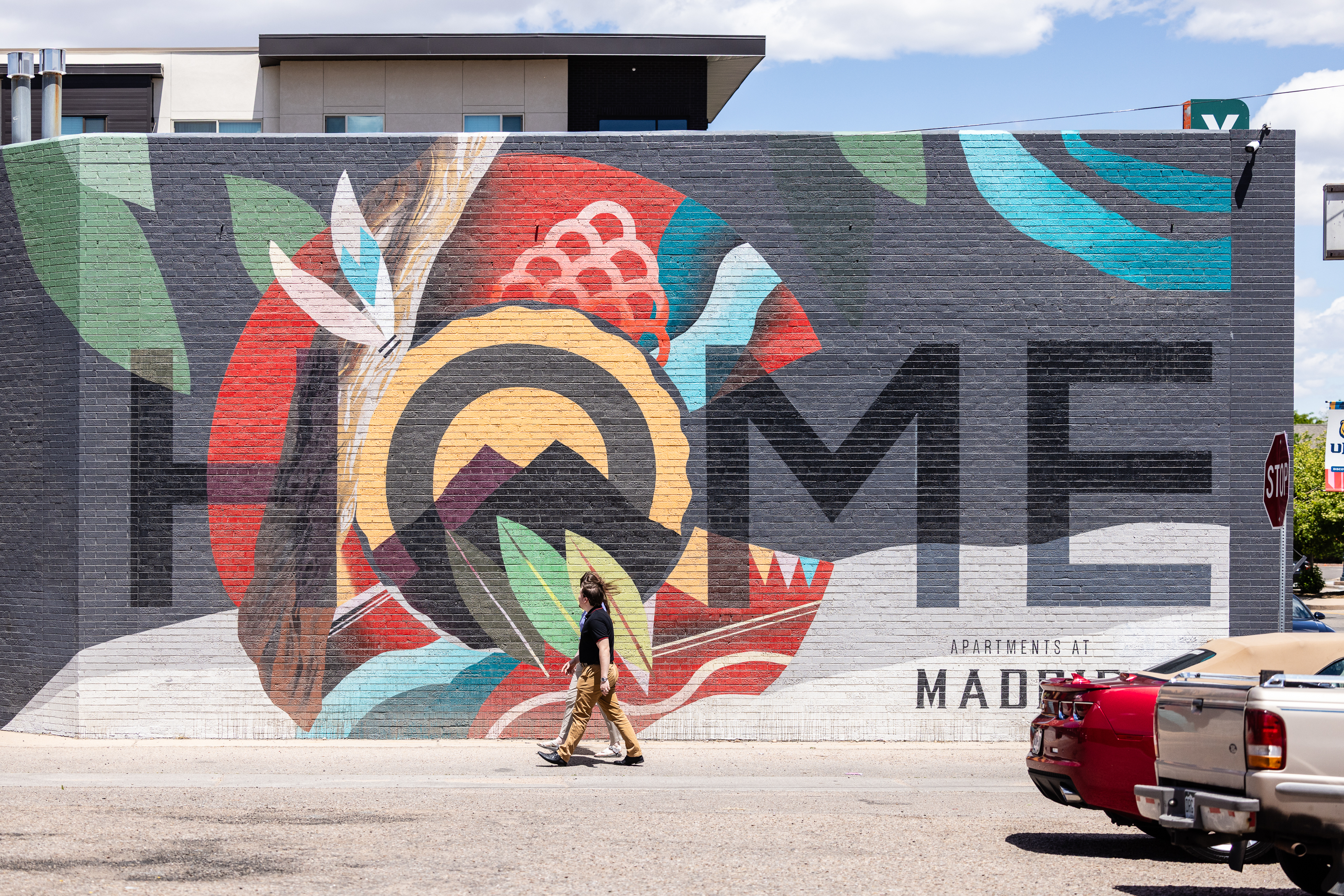 Large colorful “HOME” mural on a brick wall in Greeley, with a person walking by on a sunny day.