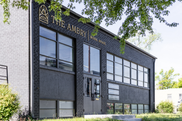This image depicts The Ambry coworking space used by Greeley startup businesses and entrepreneurs.  The building is a rectangular, two-story facility with a black brick facade and ample glass windows on both floors.  The front entrance double-doors have large glass windows above, and the building is surrounded by grass, trees, and green bushes.
