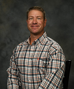 A formal portrait image featuring an individual in checkered attire seated against a neutral background.