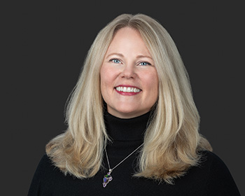 A headshot showcasing a woman with blonde hair wearing a black turtleneck and a pendant necklace.