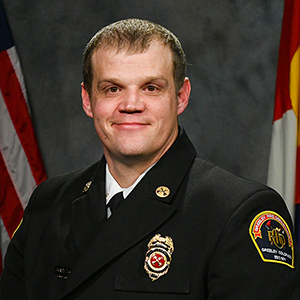 Formal portrait of a fire department official with insignia and flags in the background.