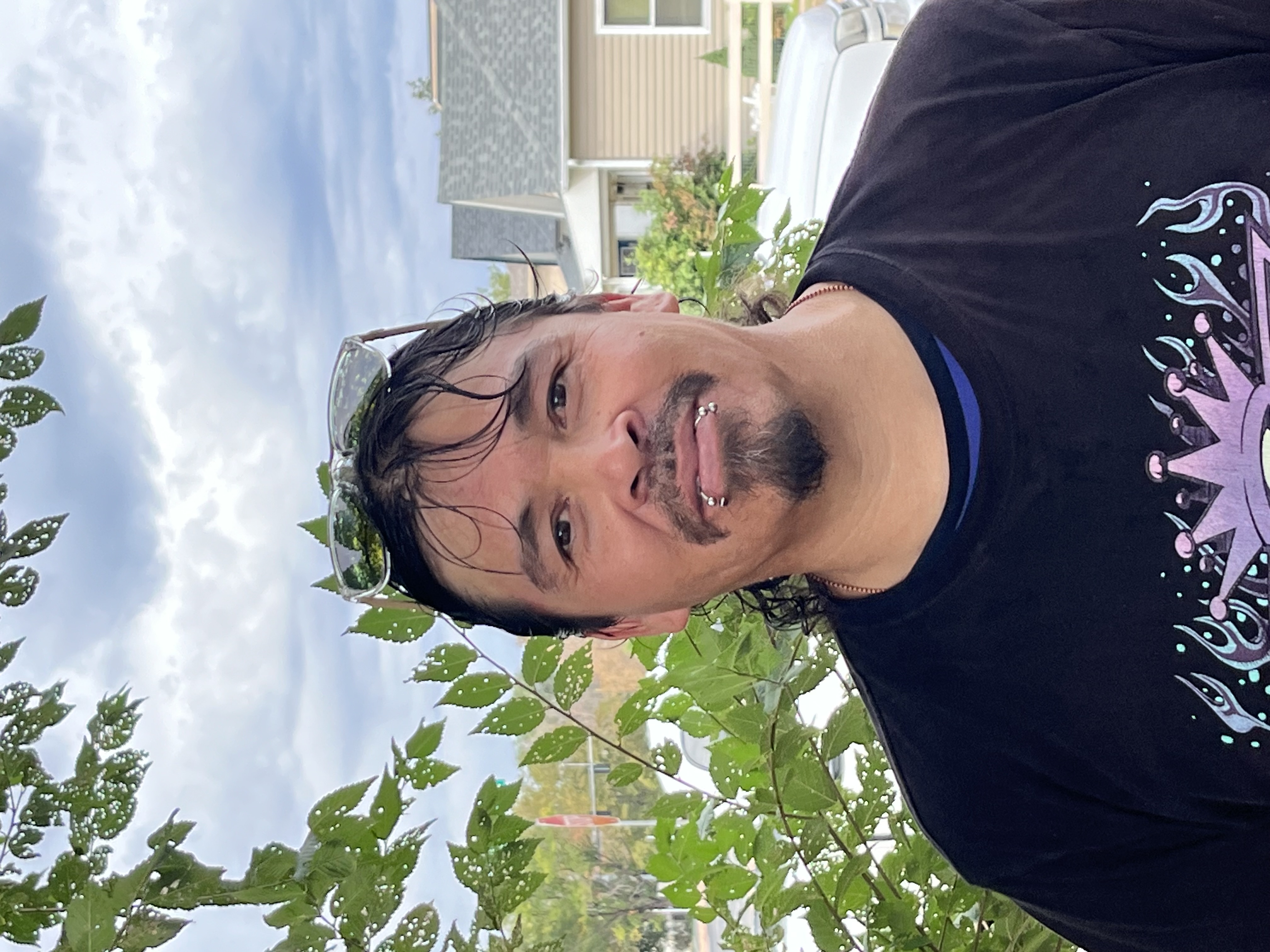 A person with black hair wearing a black shirt and sunglasses on top of head, smiling for a photo in a neighborhood. 