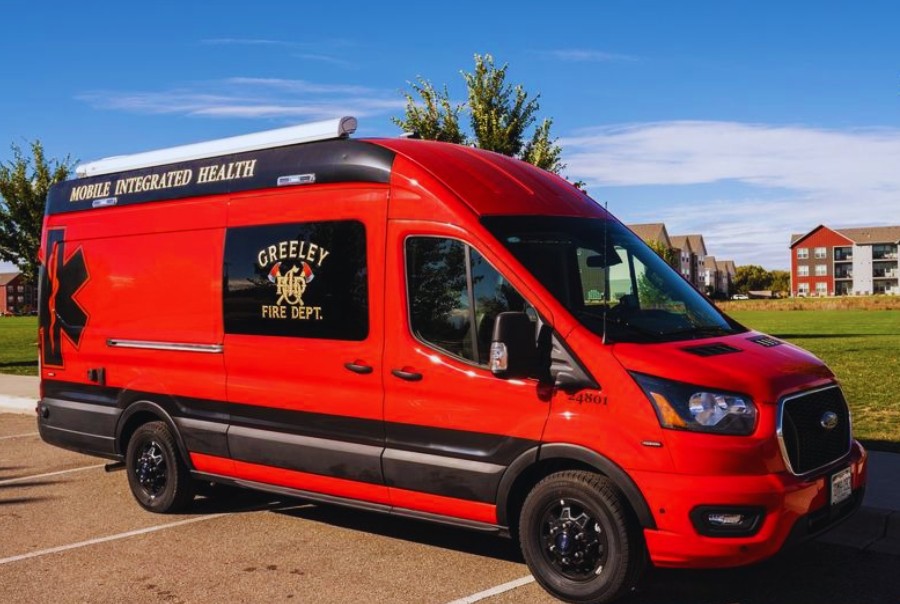 Side profile of Squad 101 Apparatus Red Sprinter Van in parking lot with buildings, grass, trees and blue skies above. 