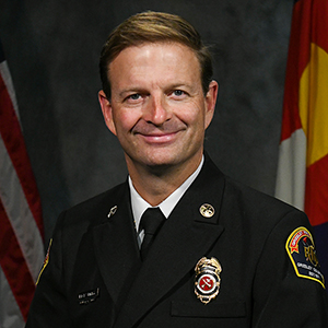 A professional portrait of Rick Smith in formal fire department uniform with badge insignias and flags in the background.