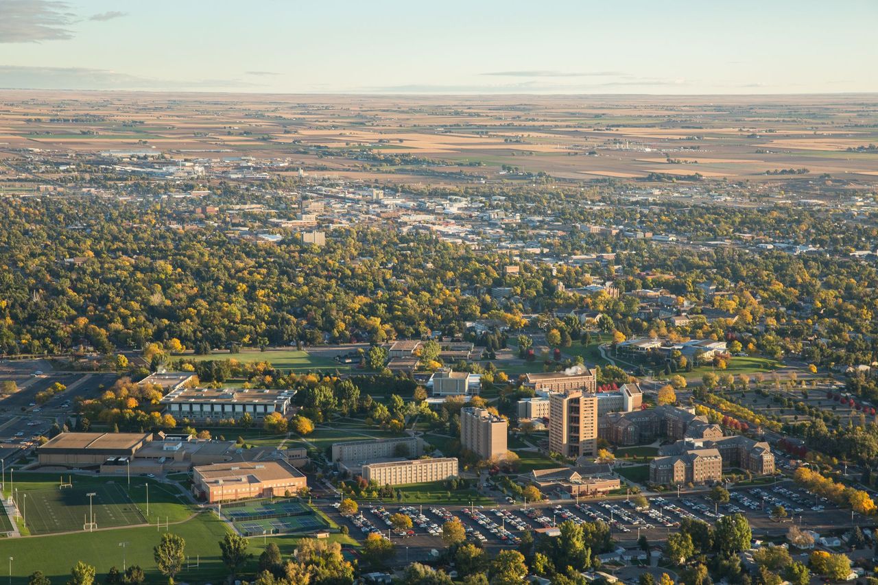 Aerial View of University of Northern Colorado (UNC) campus in Greeley, Colorado.  Expansive, red brick buildings are surrounded by trees and a grass sports field can be seen at the left edge of the image.  In the distance, behind the campus, a large expansive of trees leads to residential and commercial buildings in Greeley.