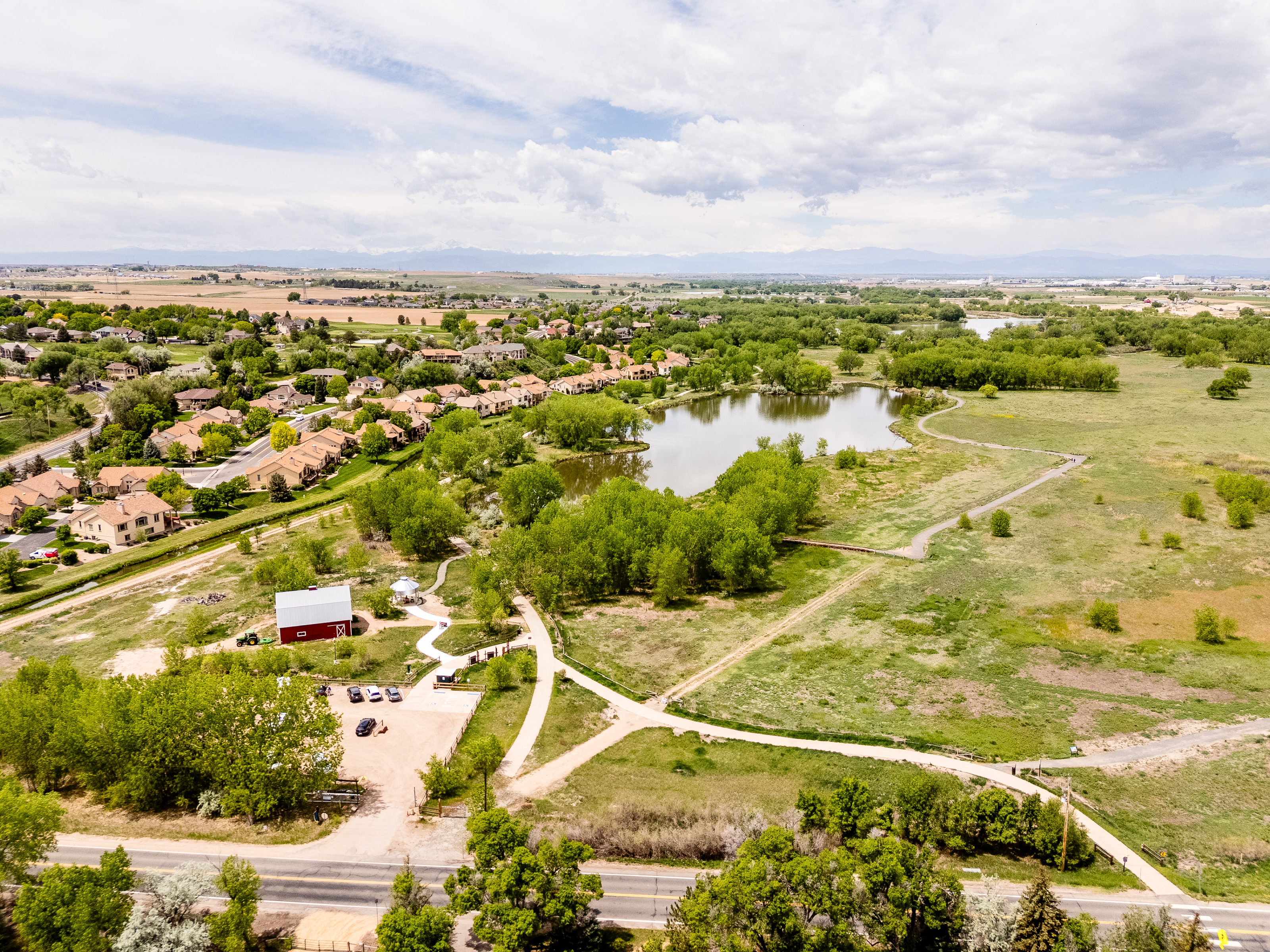 A red barn with coffered metal roof can be seen from this aerial image, with fields, trees, and a large lake behind the barn.  Paved lanes lead to the barn and around the surrounding property.  A roadway runs along the side of the property and is heavily lined with mature trees.