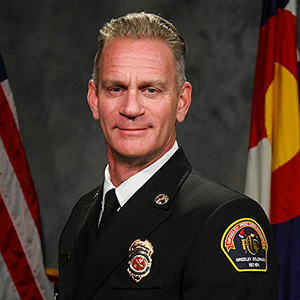Chris Mirowski in official Greeley fire uniform with American and Colorado flags.