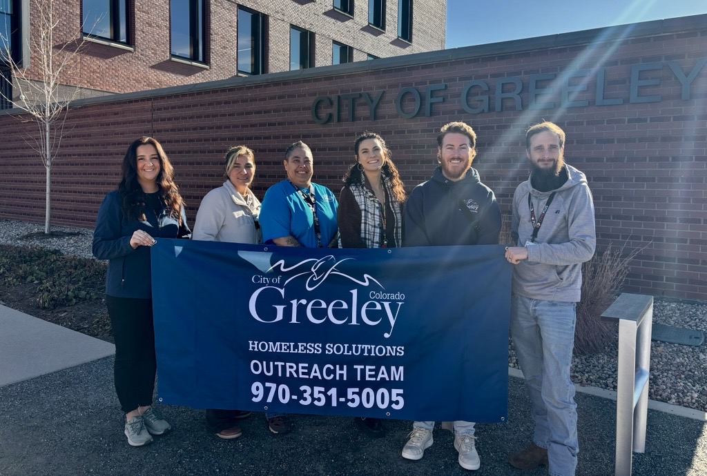 Staff members of the Homeless Solutions Outreach team outside City Center South under the City of Greeley sign holding a blue banner with the City of Greeley logo, Homeless Solutions Outreach Team, 970-351-5005. 