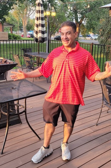 A person wearing shorts and a red shirt standing on a patio in a neighborhood smiling and holding their hands out to the side for a photo. 