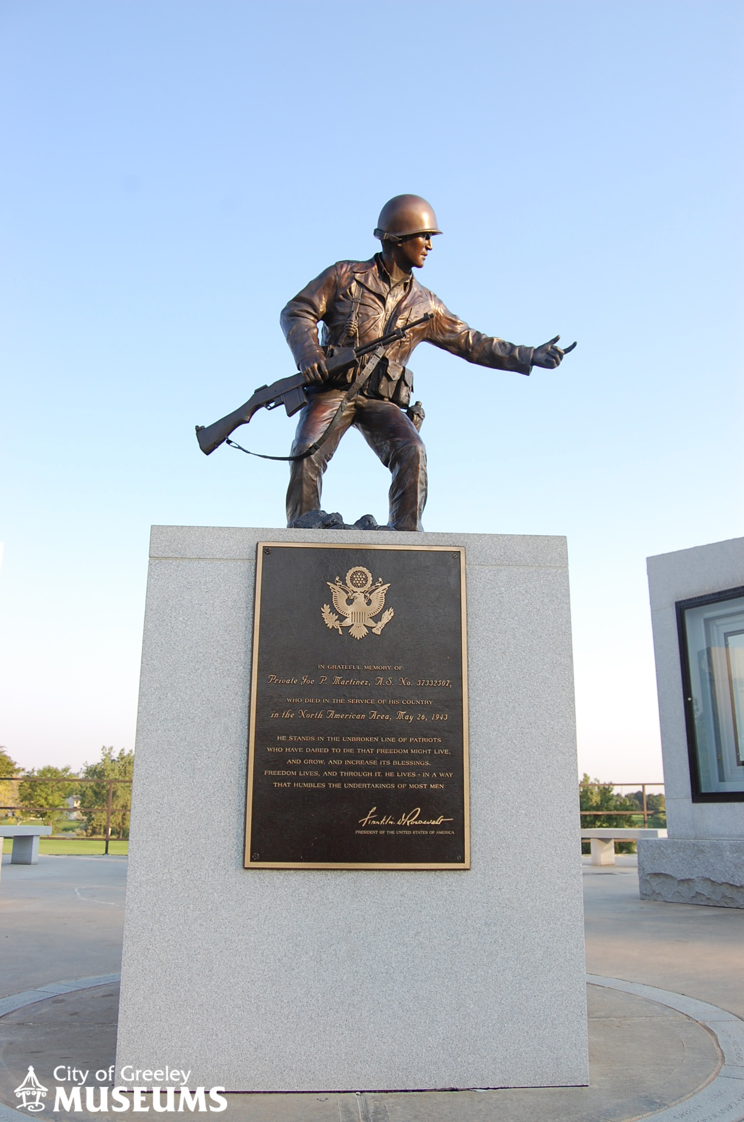 Bronze statue of a soldier wearing a uniform and helmet, carrying a gun and gesturing to his left