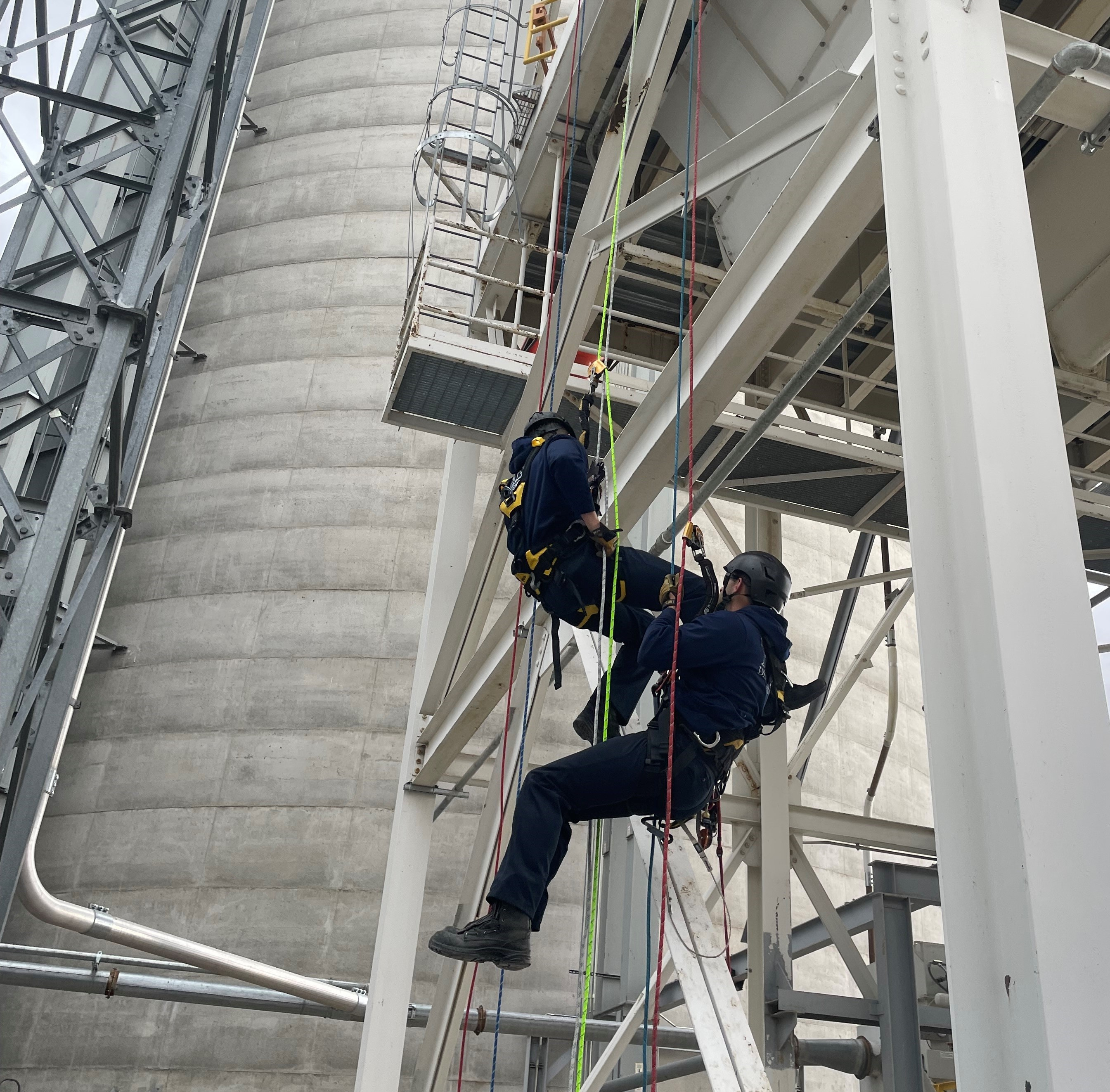 Two Greeley firefighters in blue clothing and helmets rappelling down a silo-like building with a white metal structure, using colored ropes to assist their descent.  