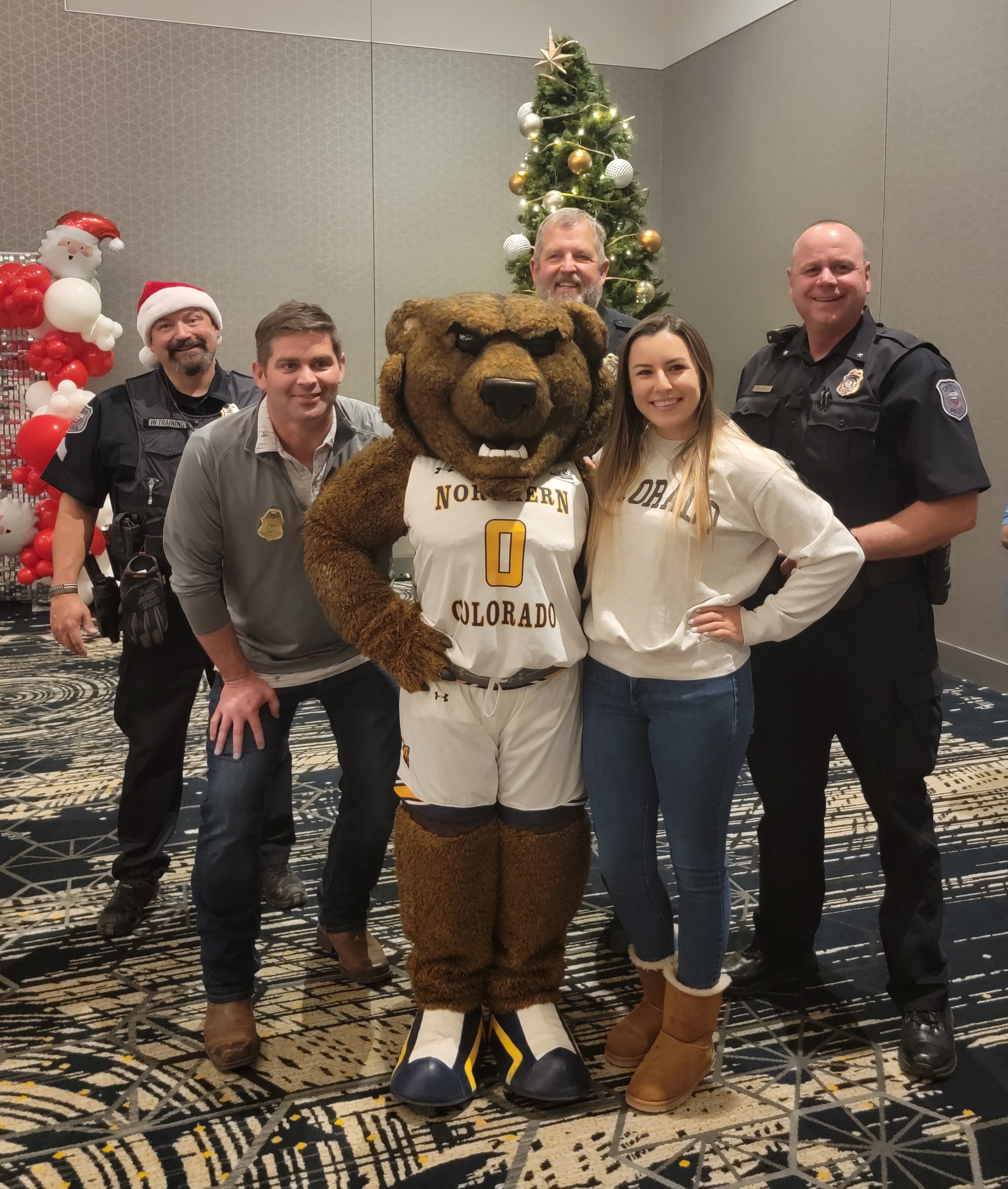 Officers with the Greeley Police Department pose with "Klawz", the brown bear mascot for the University of Northern Colorado, for a Santa Cops picture. Klawz is wearing a white basketball uniform with the number 0 on the front. There is a Christmas Tree in the background, as well as red and white balloons and a Santa balloon.