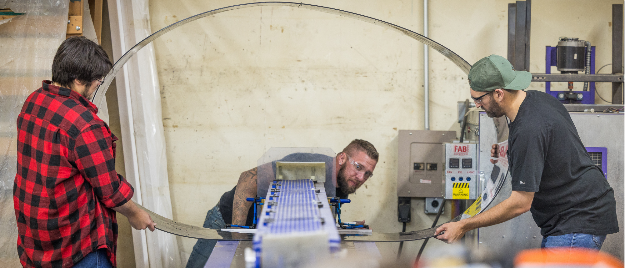 Three workers inside a manufacturing facility and working on a metal bending machine.  Two workers on each side of the machine are holding a large circular strip of metal while a third person looks on to ensure quality control.