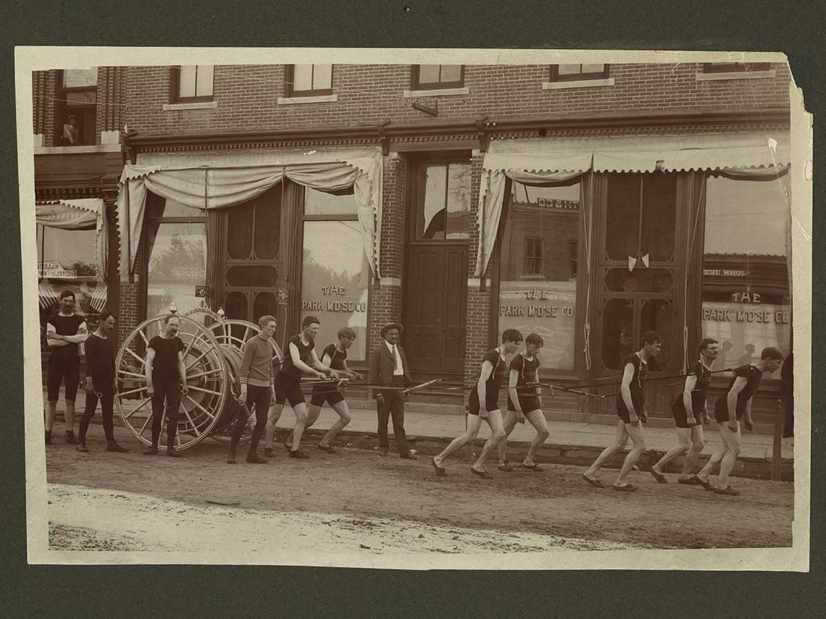 Historic photo circa 1988 of a group of young men hitched to a large hose reel on the street in front of brick storefronts labelled The Park MDSE CO
