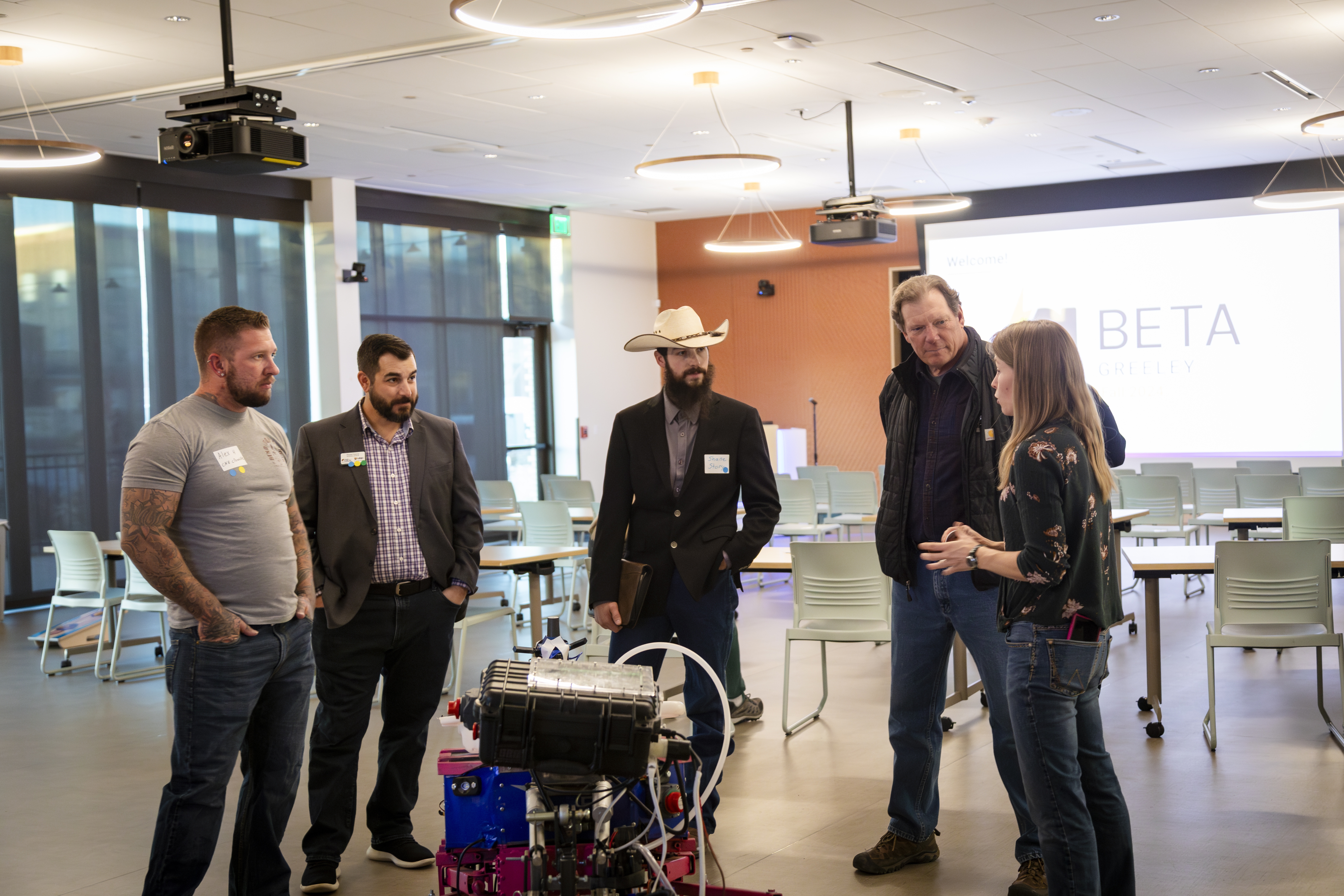 A group of five people stand in a modern meeting room having a discussion near a mechanical device or engine prototype. The group includes three men in business-casual attire, one wearing a cowboy hat, and a woman explaining something while gesturing toward the equipment. A presentation screen in the background displays the logo “ALPHA BETA Greeley.”