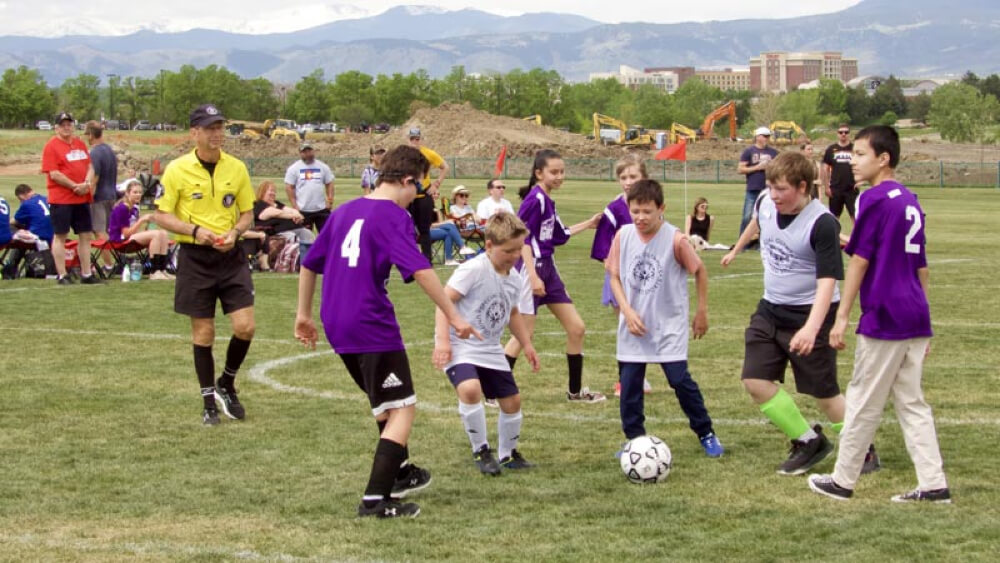 Four youth unified soccer players compete against each other while the referee looks on during a game outside on a grassy field overlooking the mountains