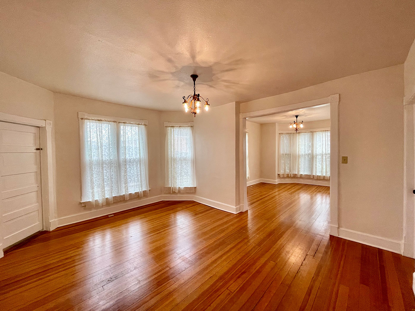 Interior view of two parlors connected by a doorway. The rooms have wooden floors, white walls, and small light fixtures.