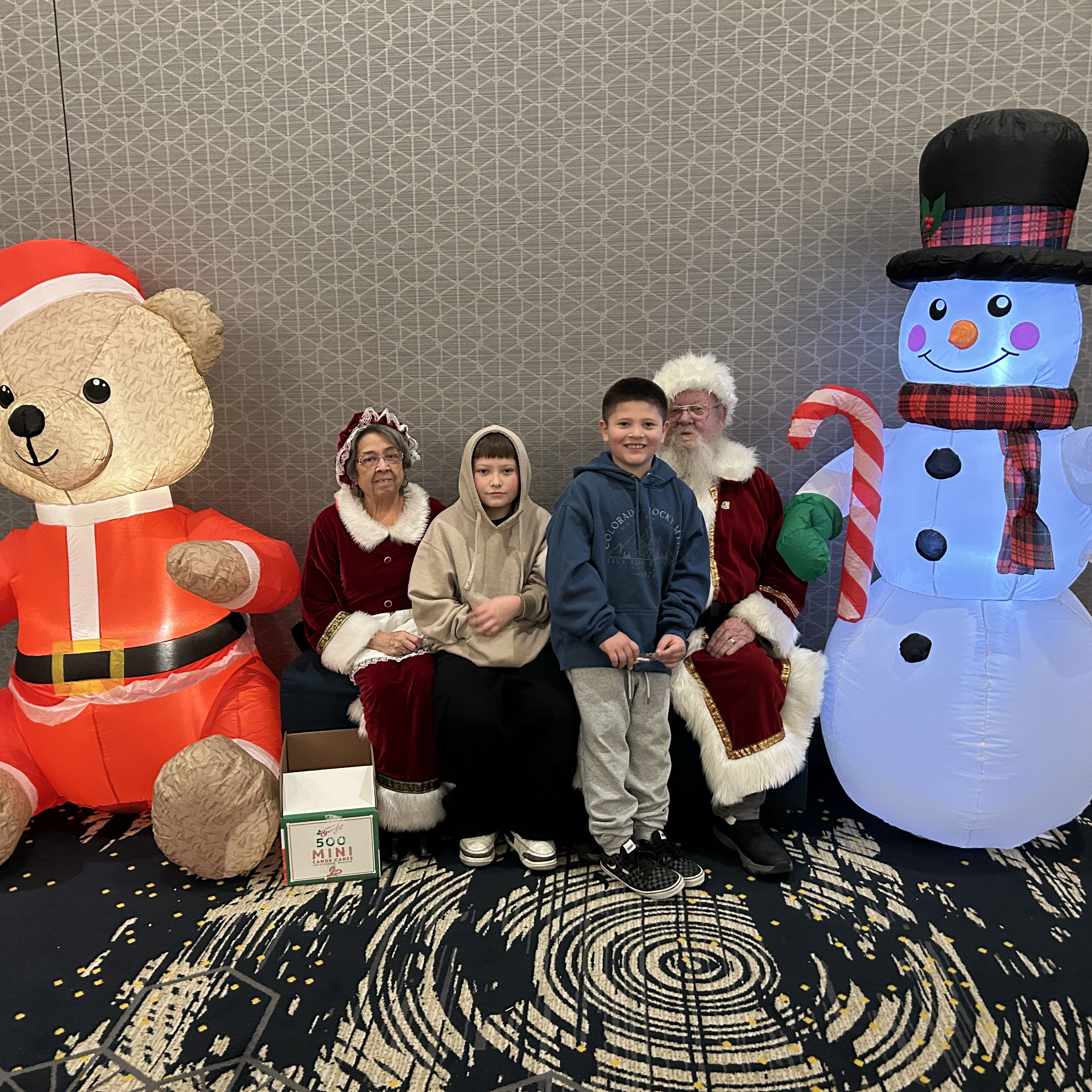 Two little boys pose for a photo between Mrs. Claus and Santa Claus at a Greeley Police Department party. An inflatable teddy bear wearing a Santa costume is next to Mrs. Claus and an inflatable snowman wearing a top hat and holding a giant candycane is next to Santa.