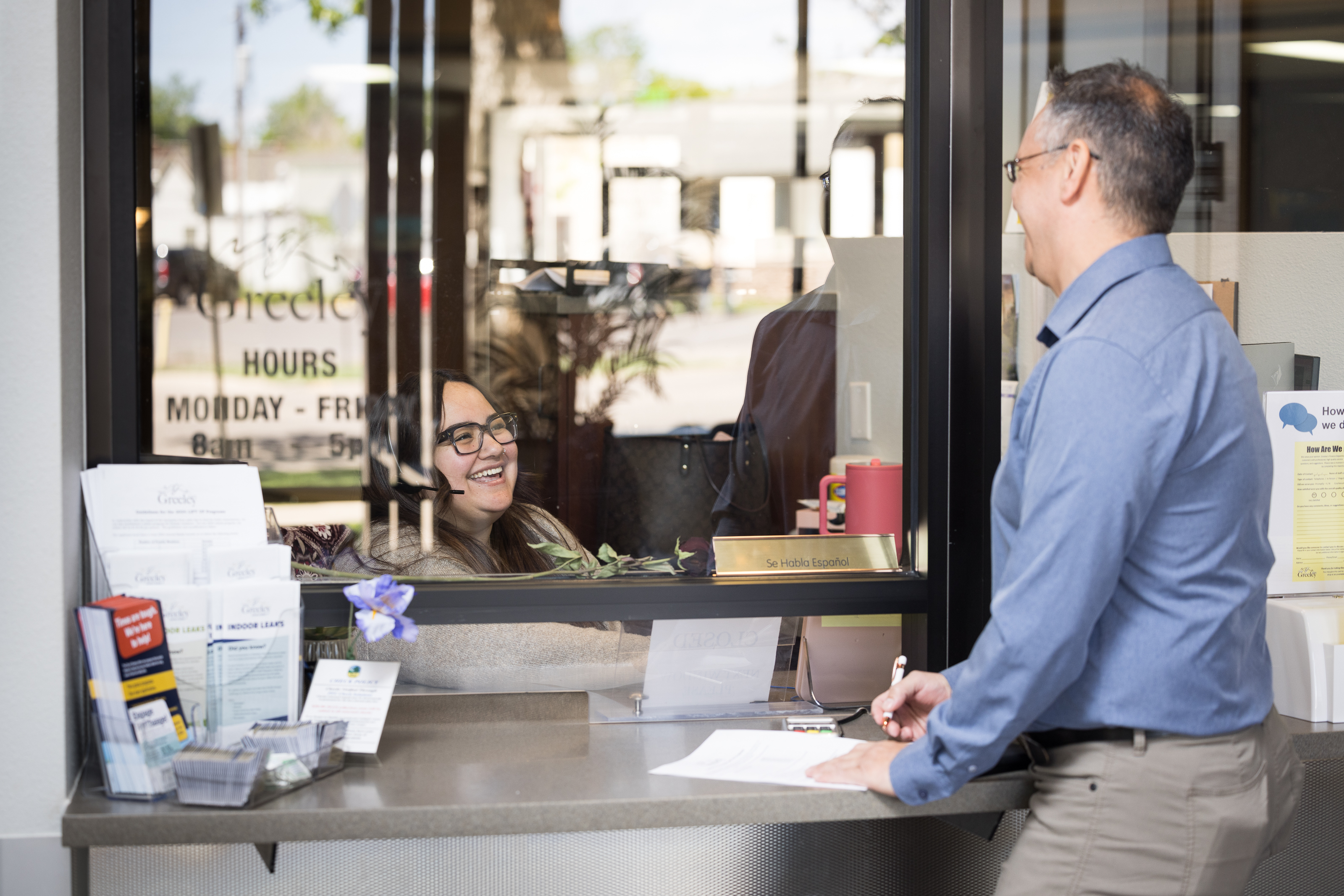 A man stands at a service window inside City Hall, speaking with a smiling City of Greeley clerk who is sitting behind the glass. Brochures and informational pamphlets are displayed on the counter, and a sign on the window lists office hours.